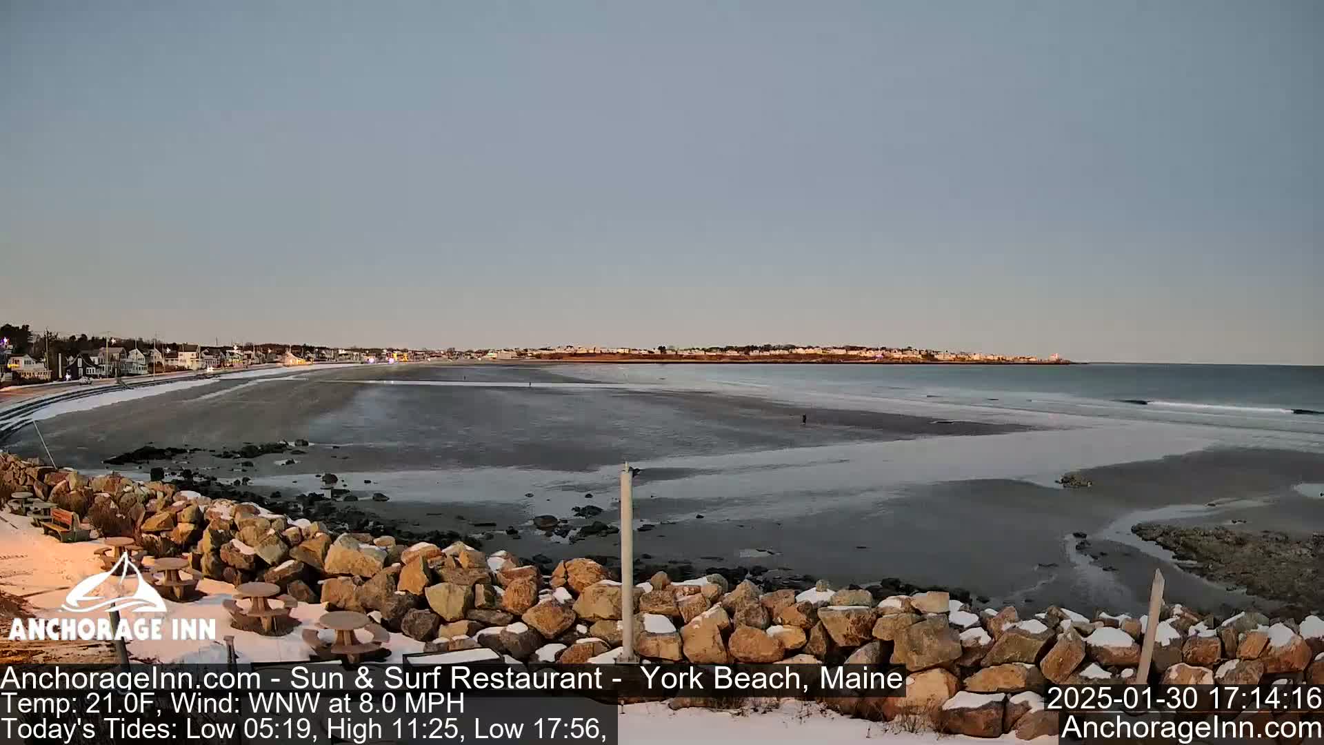 A snow-dusted rocky shoreline overlooks a partially frozen beach and calm ocean under a twilight sky.