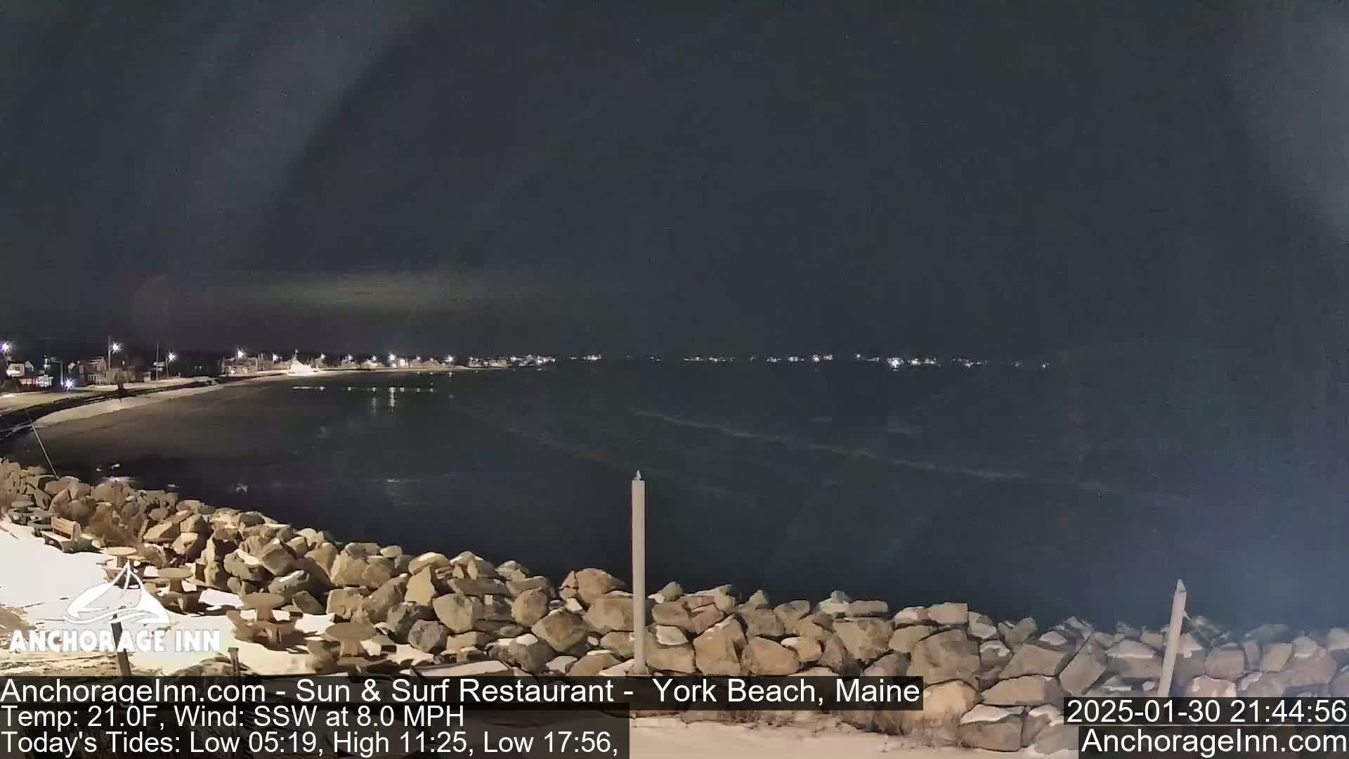 A nighttime view of a dark, rocky shoreline and calm ocean, with a line of lights visible in the distance under a clear sky.