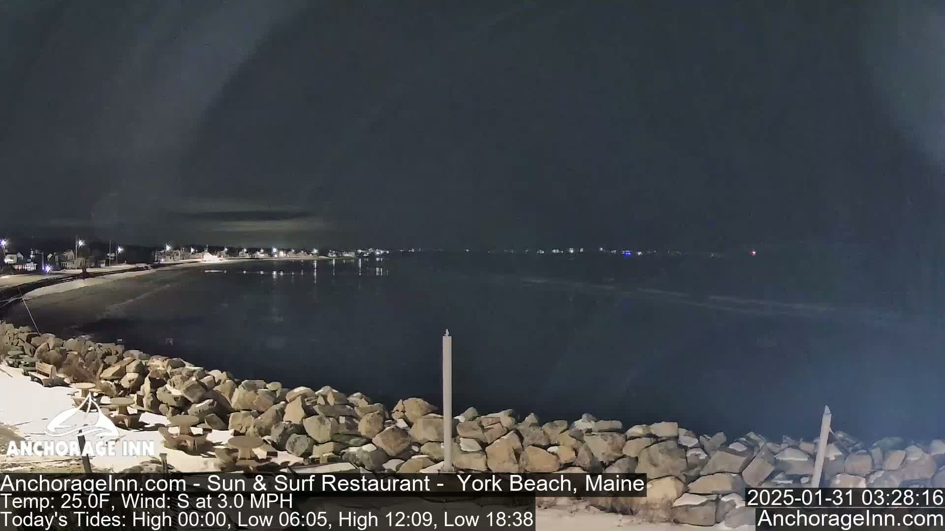 A nighttime view of a rocky shoreline and calm ocean water, with a small town's lights visible in the distance under a dark sky.