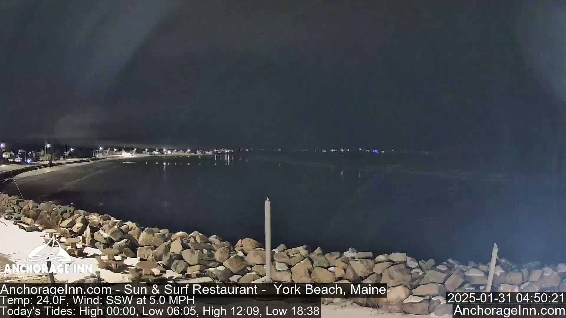 A nighttime view of a rocky shoreline and calm dark water, with a town faintly visible in the distance under a dark sky.