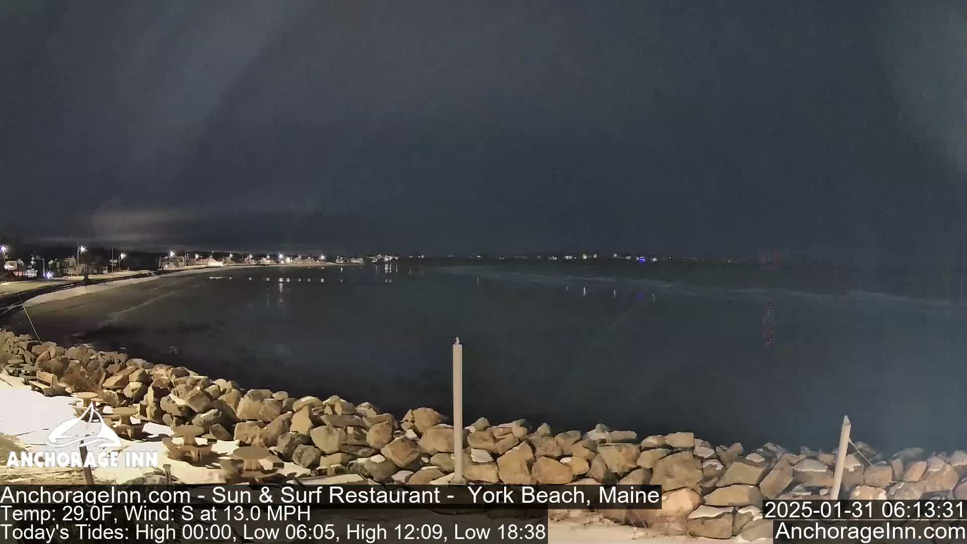 A dark, nighttime view of a rocky shoreline and calm water, with a beach and distant lights visible under a mostly clear sky.