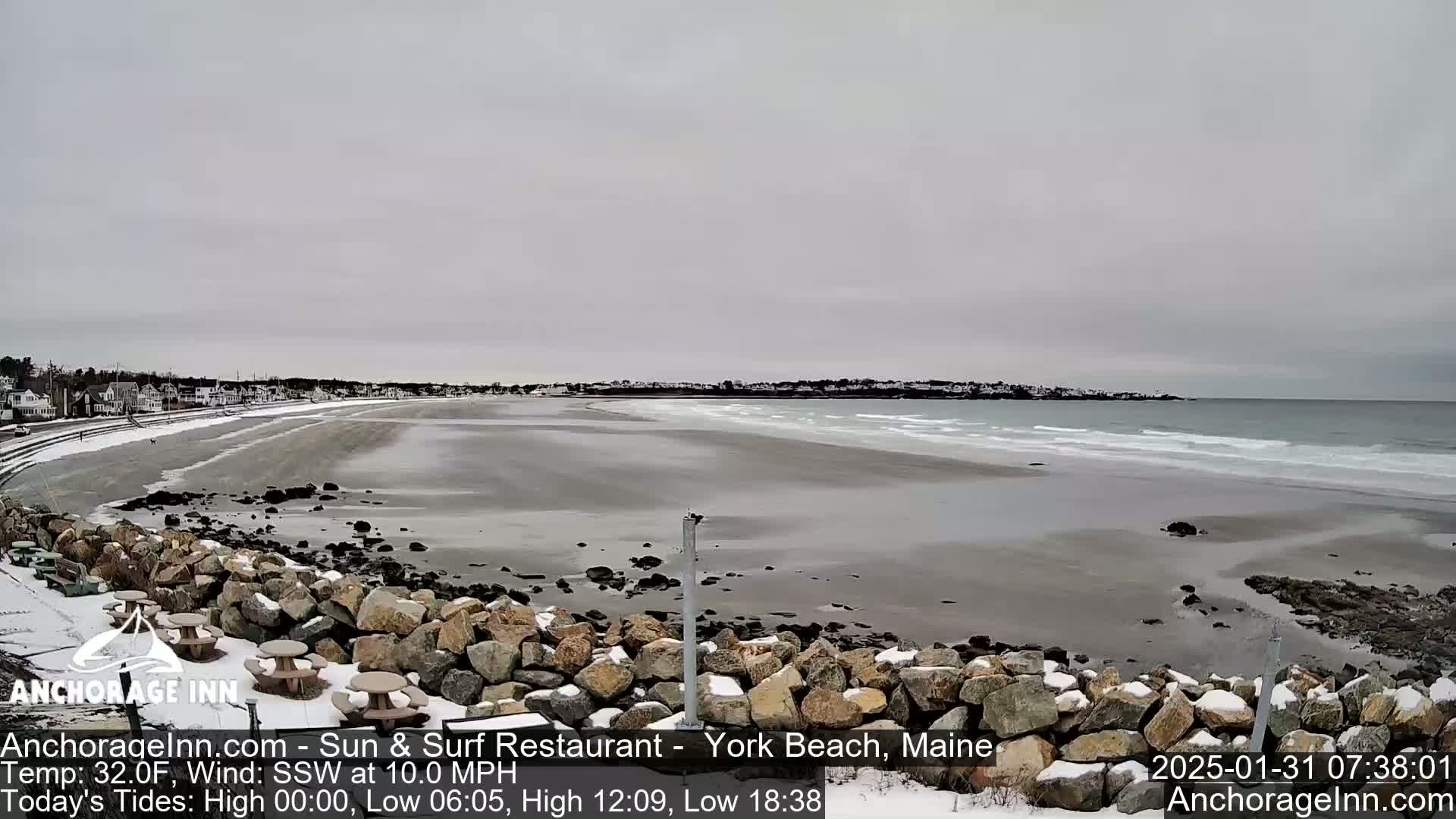 A snow-dusted rocky shoreline overlooks a wide, mostly calm beach and ocean under an overcast sky.