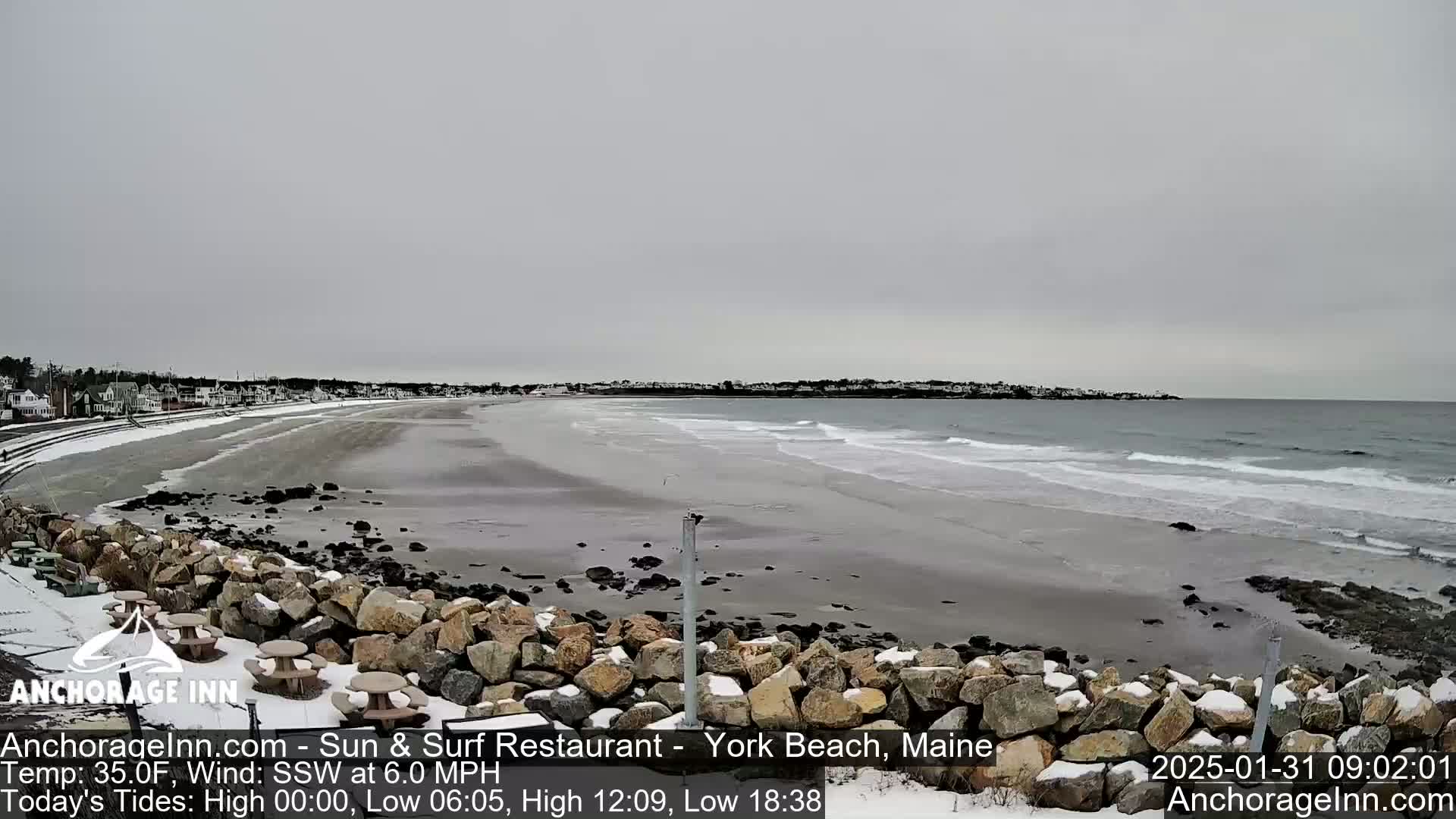 A snow-dusted rocky shoreline and sandy beach meet a gray, overcast ocean with small waves.
