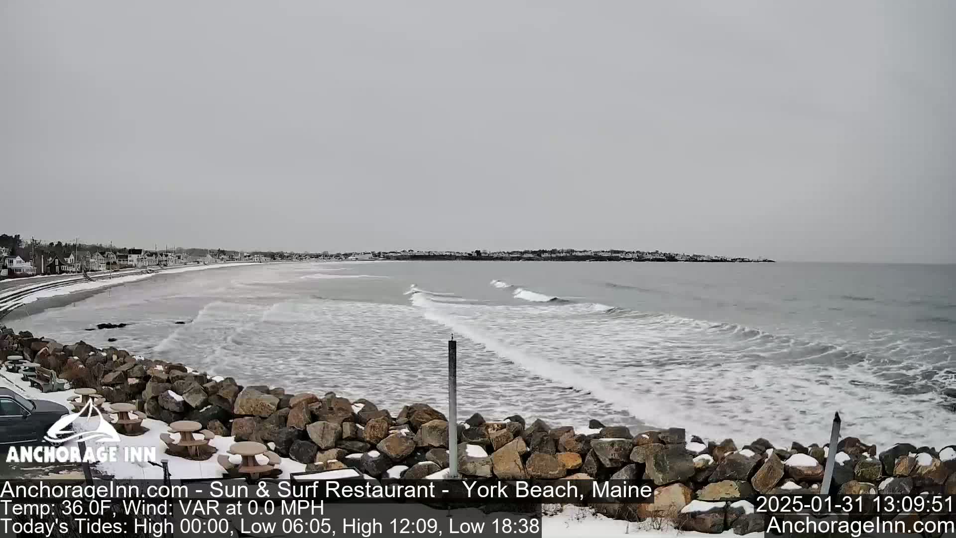 A snow-covered beach with small waves and a calm ocean under an overcast sky.