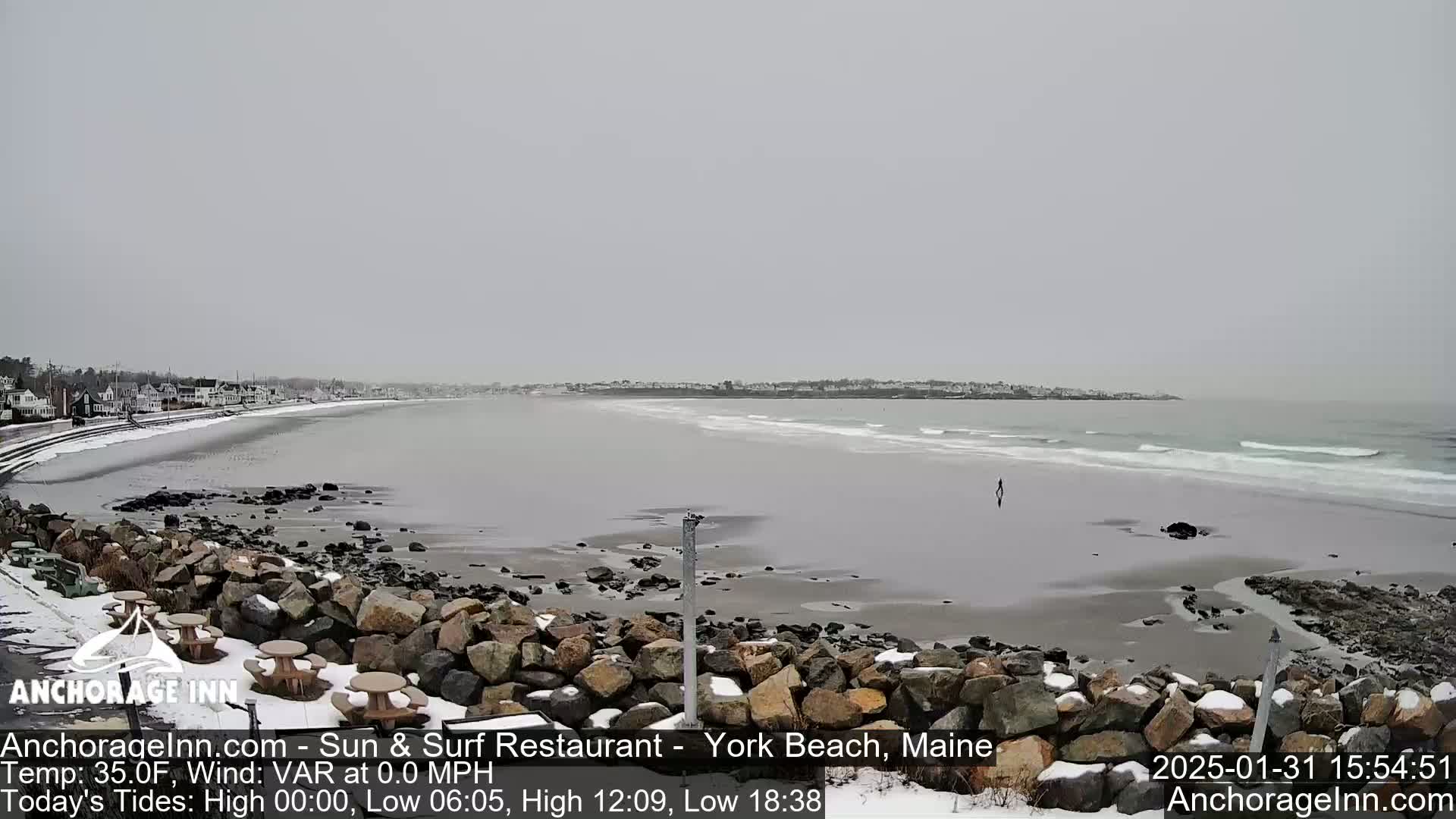 A snow-dusted beach with calm waves and a person walking in the distance, under an overcast sky.