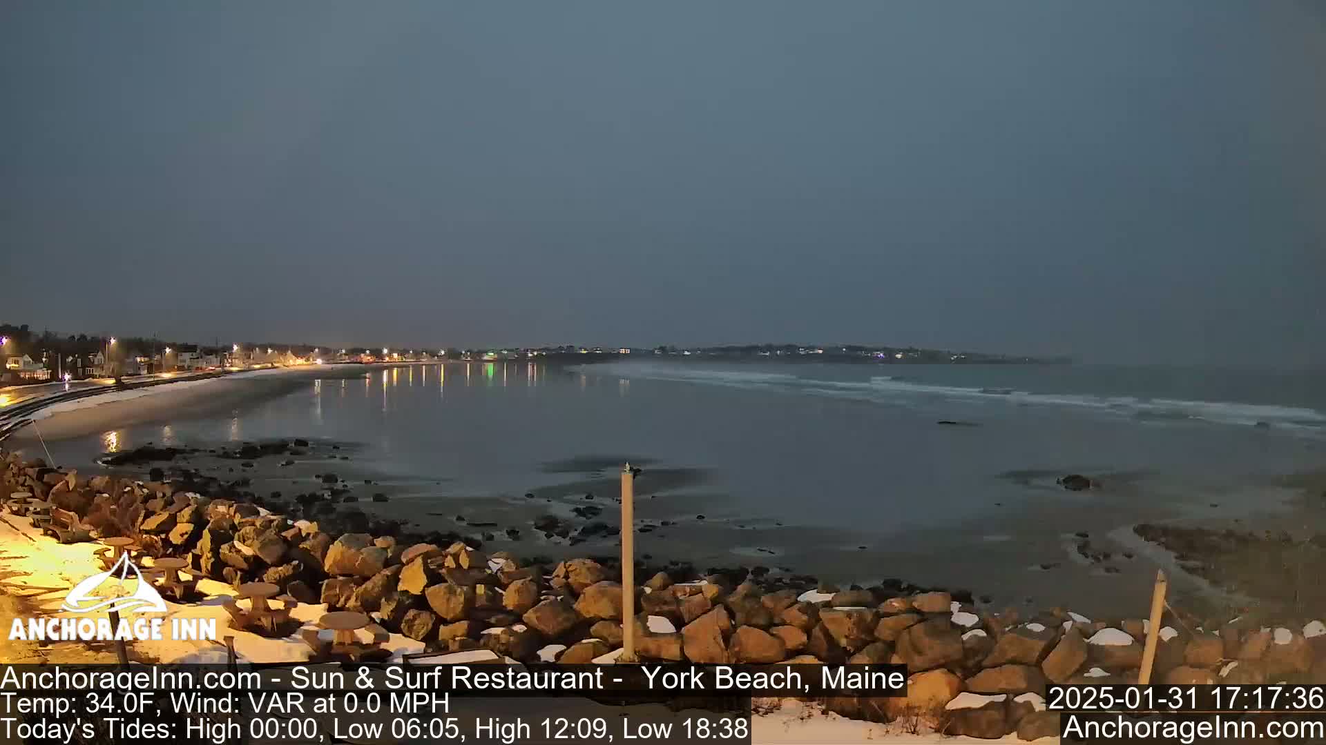 A calm, dark ocean with small waves laps a snow-dusted rocky shoreline at twilight, under a gray sky.
