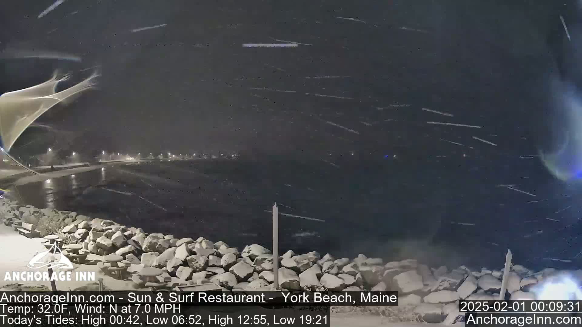A nighttime scene of a snow-covered rocky shoreline and dark water under a heavy snowfall.
