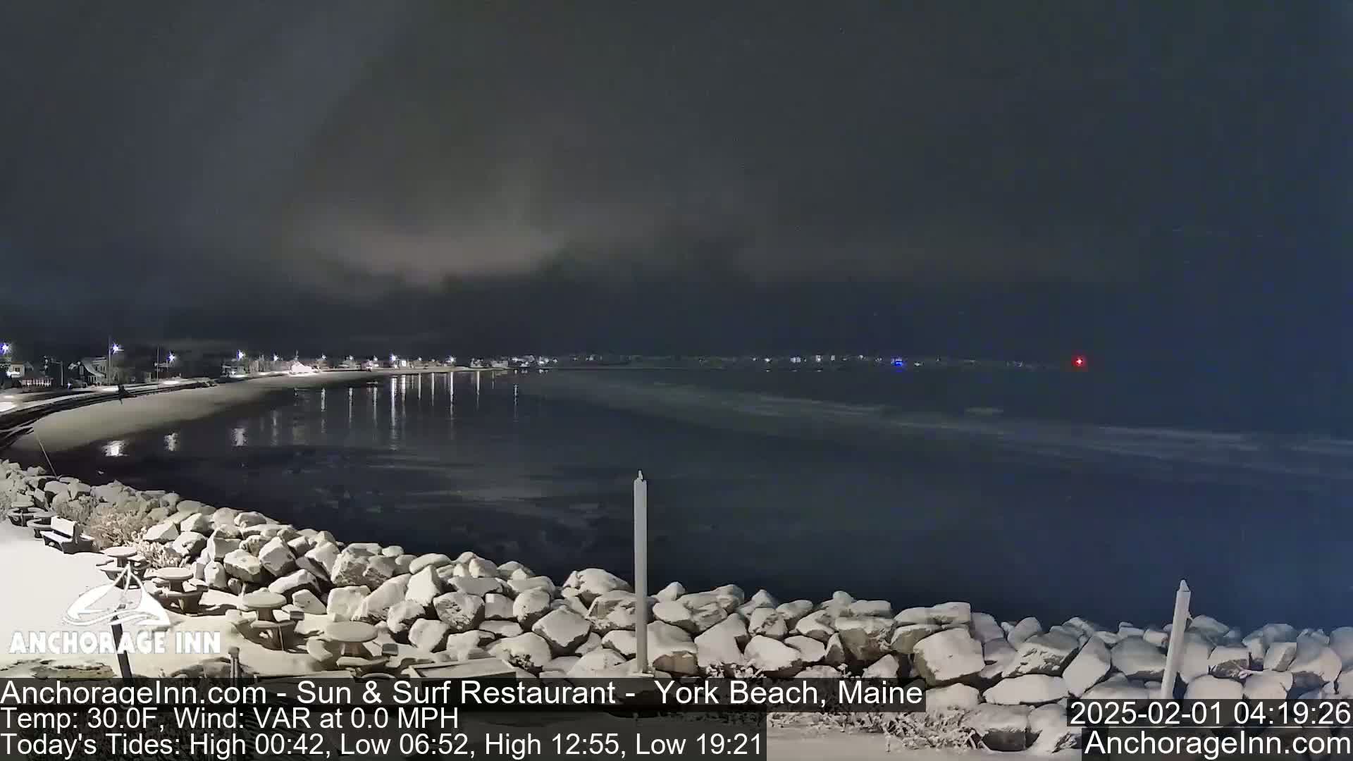 A snow-covered rocky shoreline at night overlooks a calm, dark body of water with a town faintly lit in the distance under a cloudy sky.