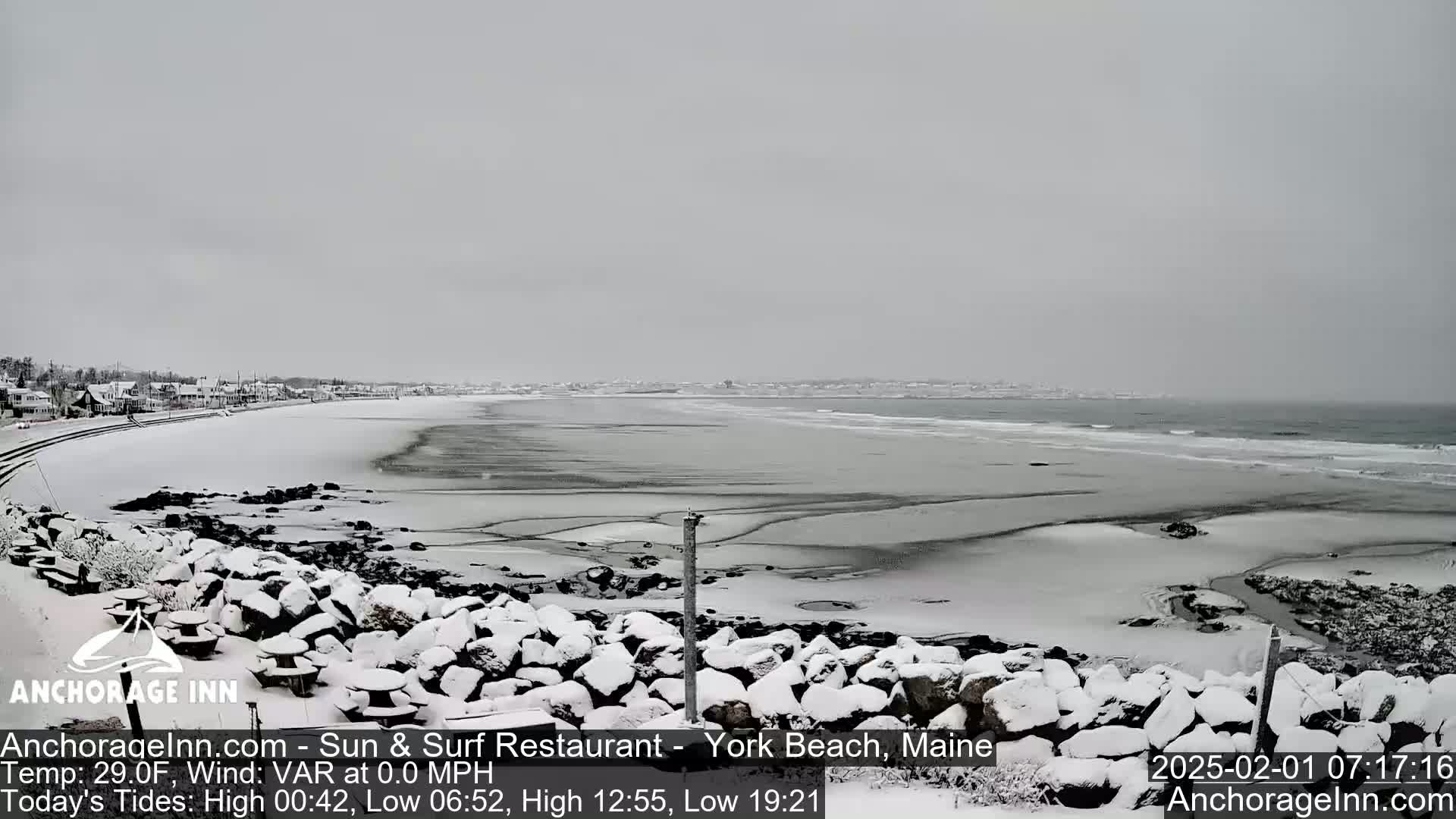 A snow-covered beach scene shows a calm ocean with a shoreline of rocks and a distant town under an overcast sky.