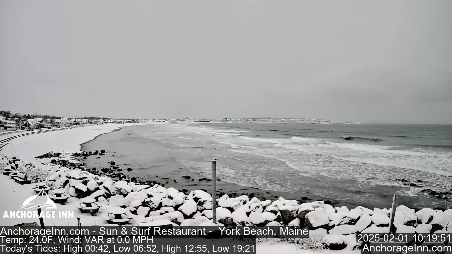 A snow-covered rocky shoreline and beach meet a gray, wavy ocean under an overcast sky.