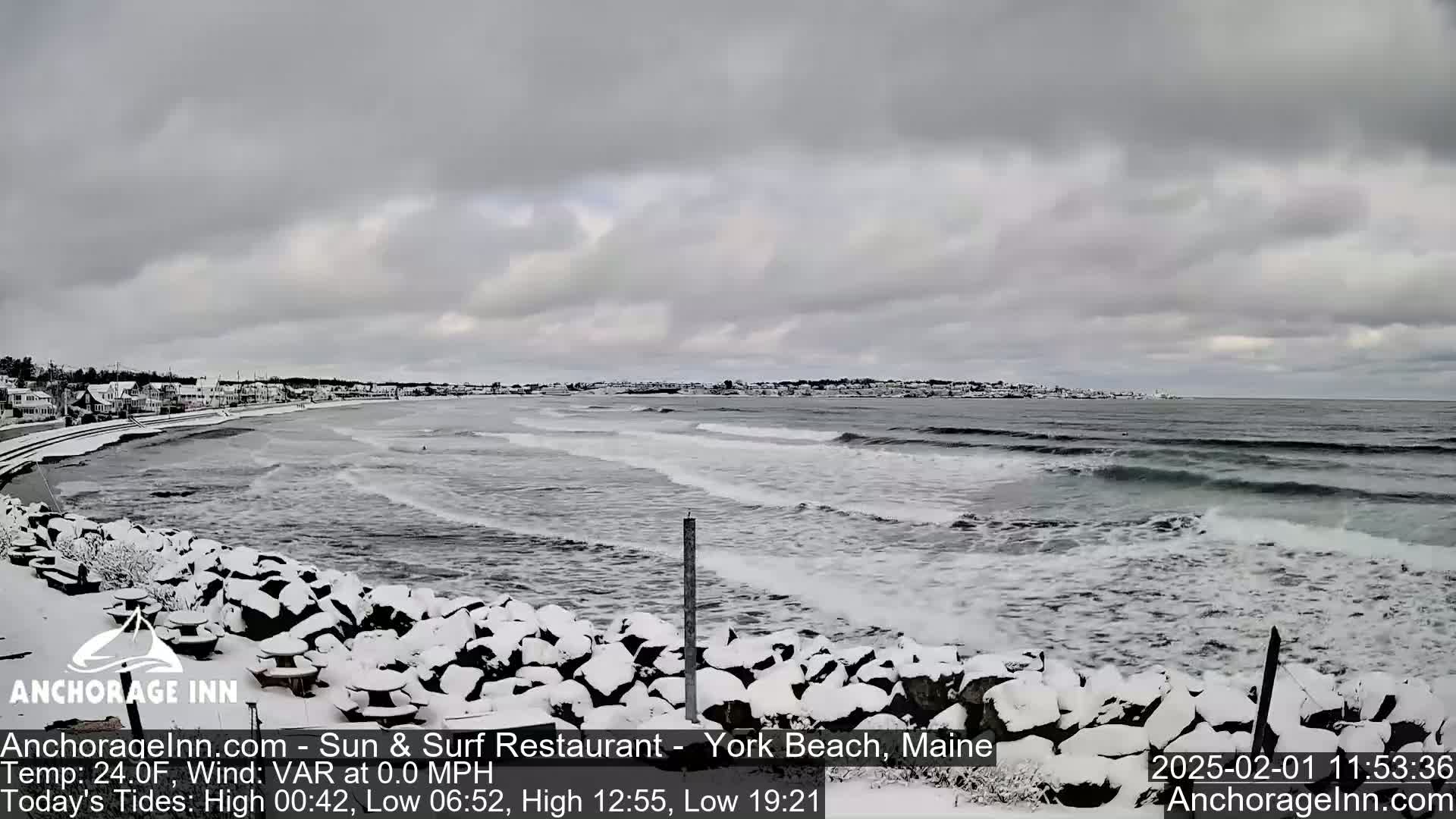 A snow-covered rocky shoreline with picnic tables overlooks a gray, choppy ocean under a cloudy sky.