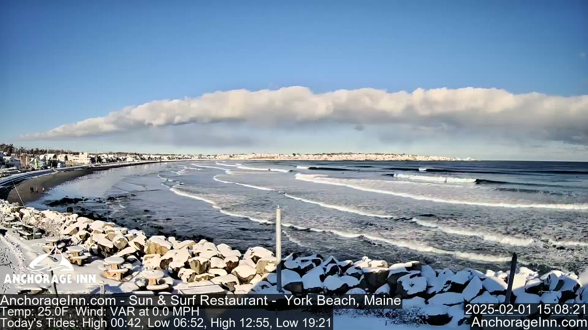 A snow-covered rocky shoreline overlooks a beach with small waves rolling in under a partly cloudy sky.