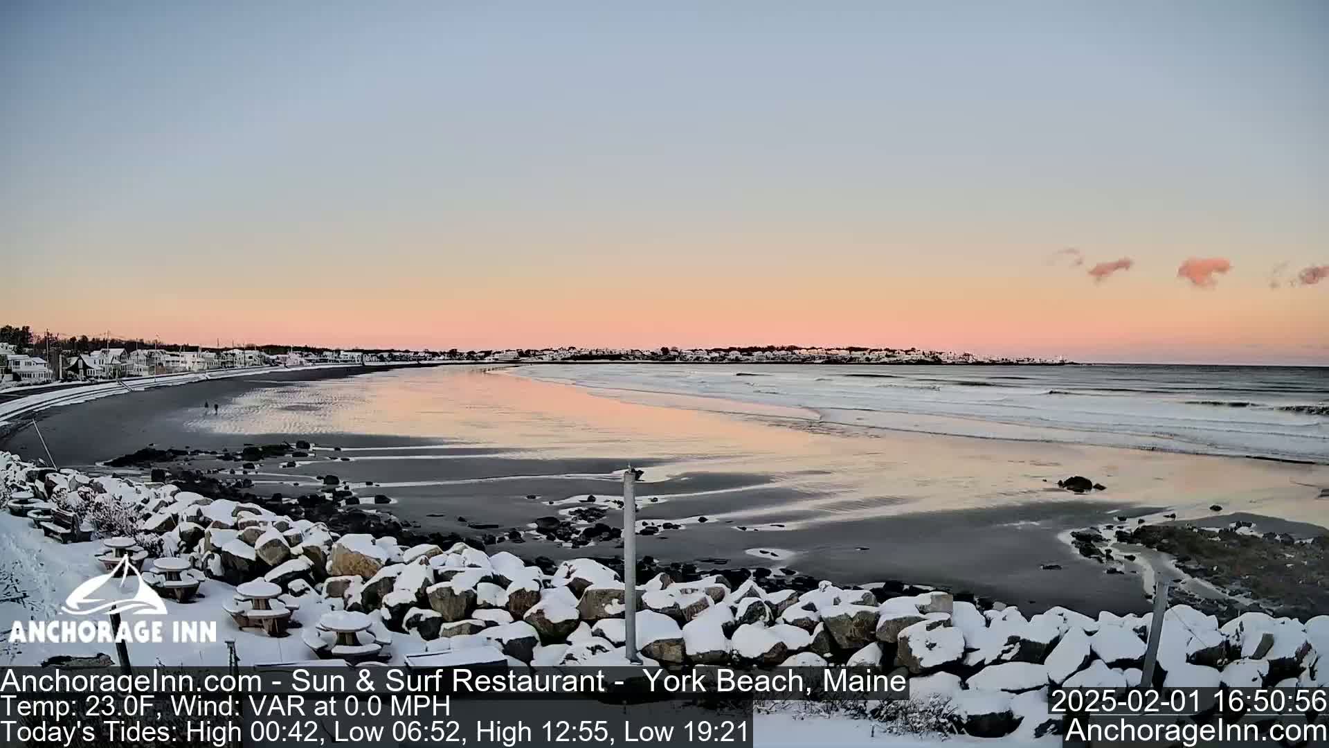 A snow-dusted rocky shoreline faces a calm ocean at sunset, with a town visible in the background.
