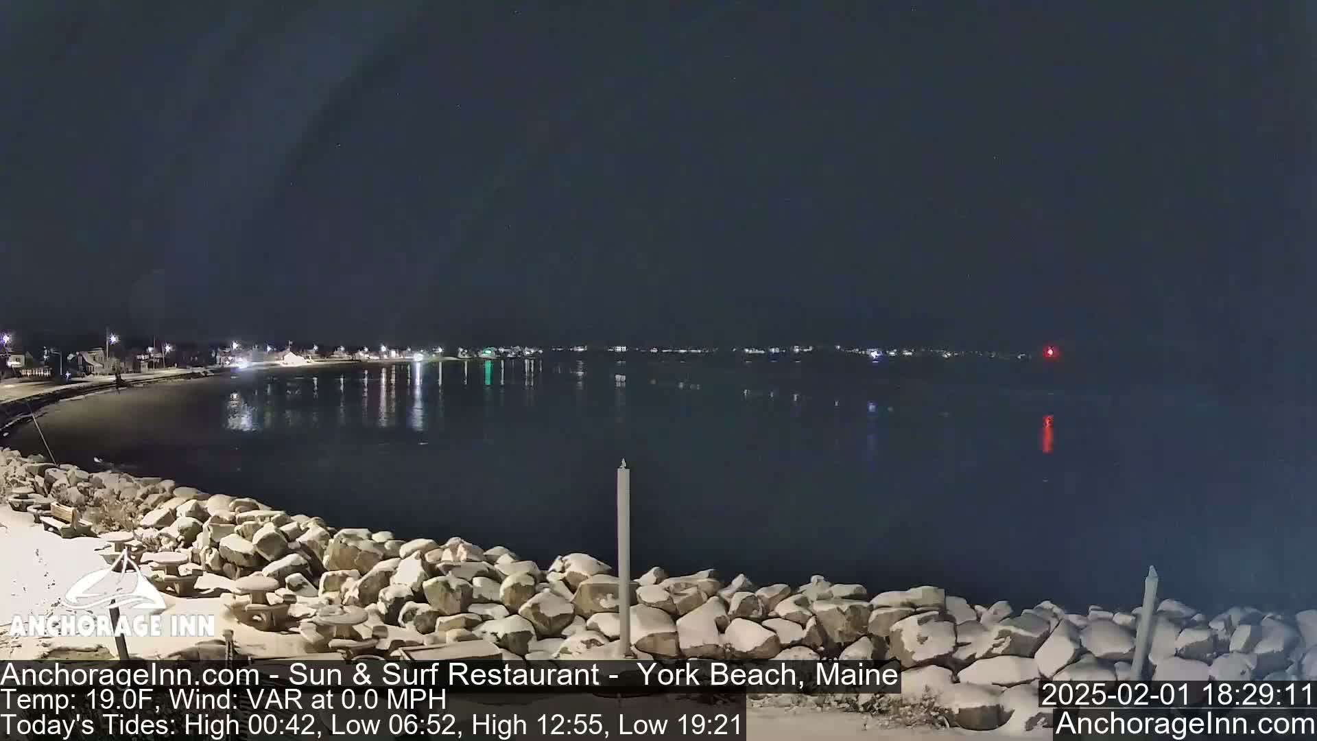 A snow-dusted rocky shoreline at night overlooks a calm, dark body of water reflecting the lights of a distant town under a clear sky.