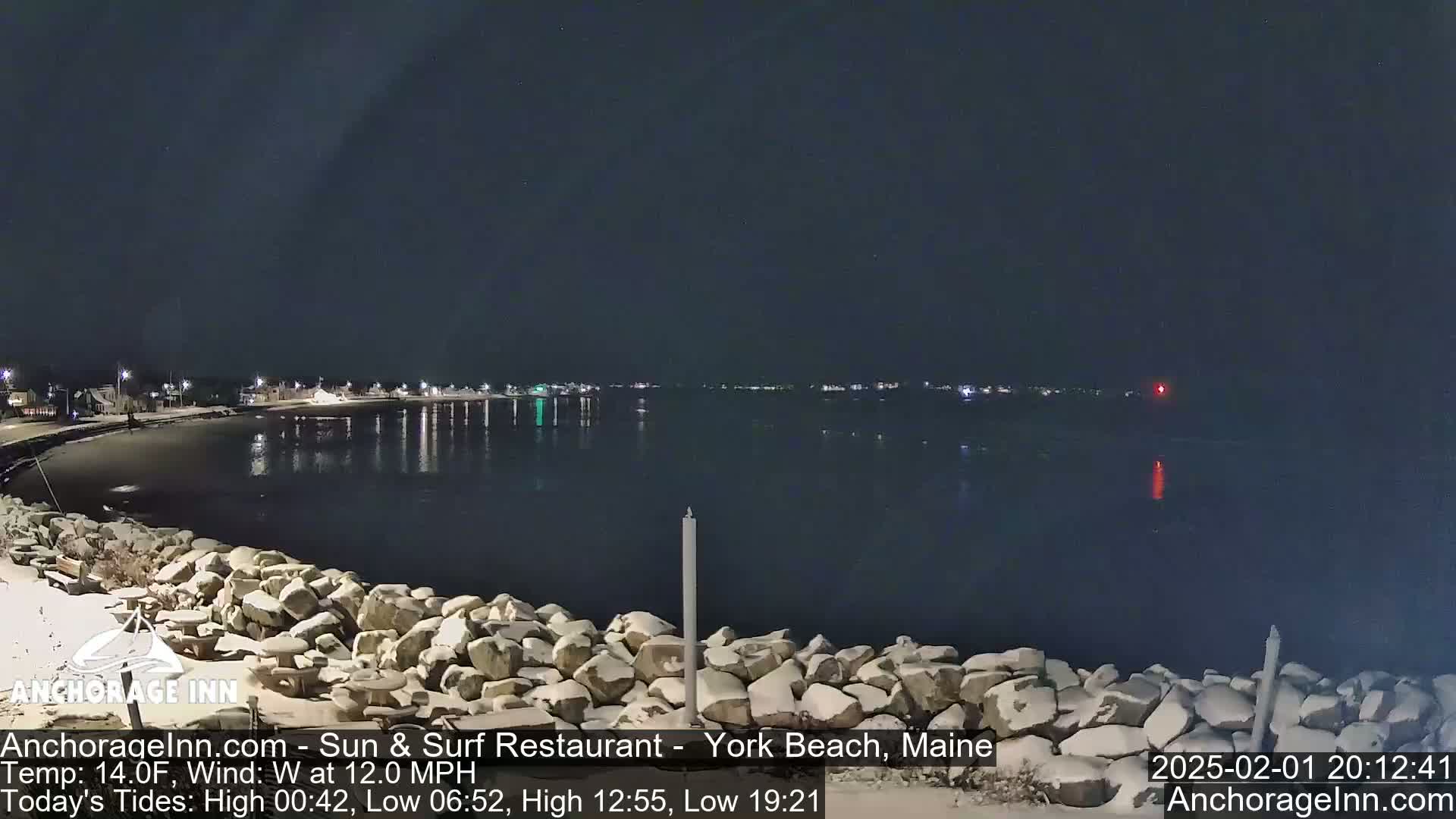A nighttime view of a snow-dusted rocky shoreline and calm water, with a town's lights faintly visible across the water under a clear, dark sky.