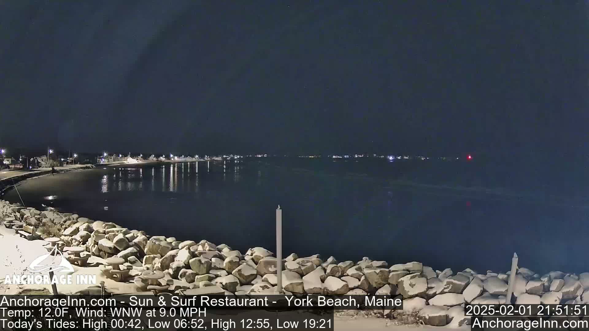 A snow-dusted rocky shoreline at night overlooks a calm, dark body of water with faintly visible lights across the far shore under a clear, dark sky.