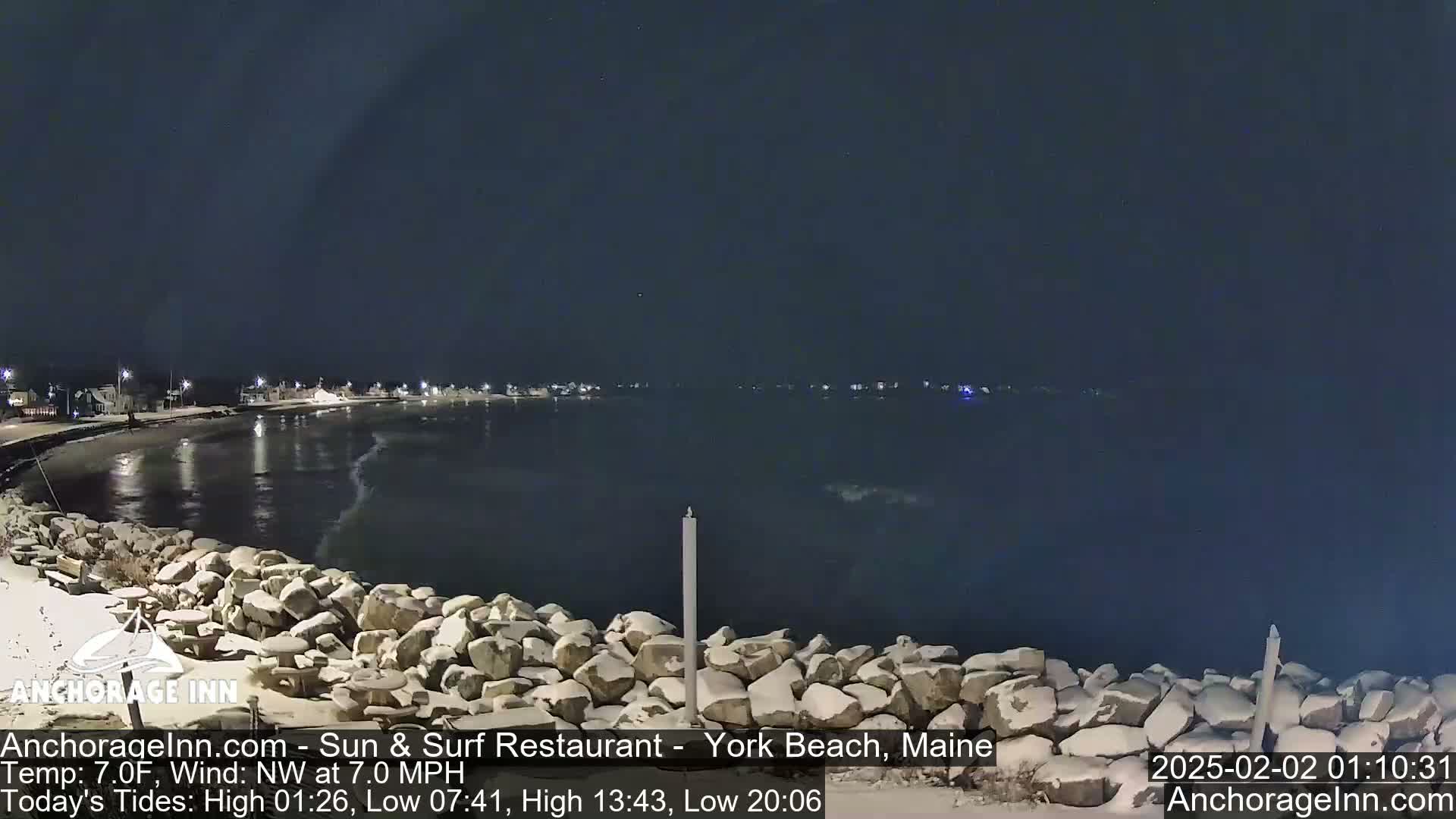 A nighttime view of a snow-dusted rocky shoreline and calm dark water, under a clear, dark sky.