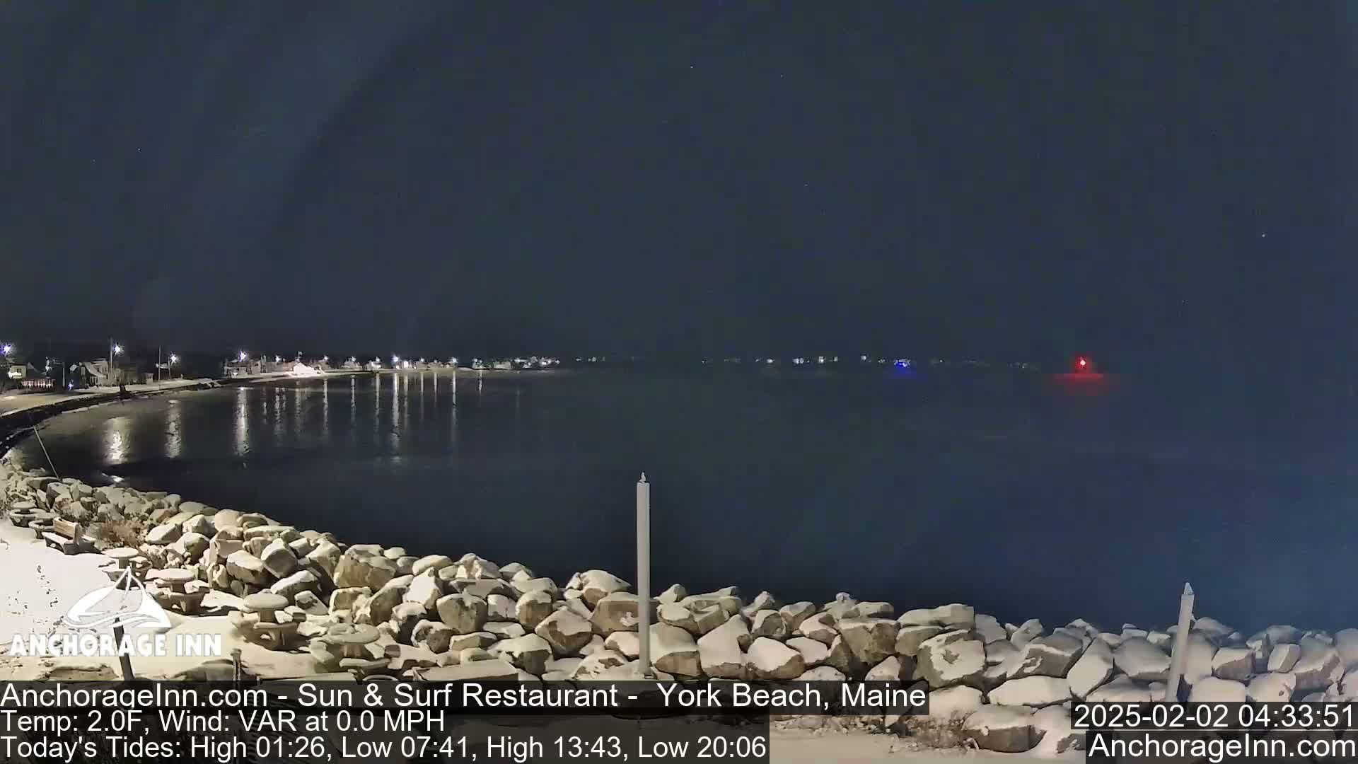 A snow-covered rocky shoreline at night overlooks a calm, dark body of water with a few distant lights visible across the water.