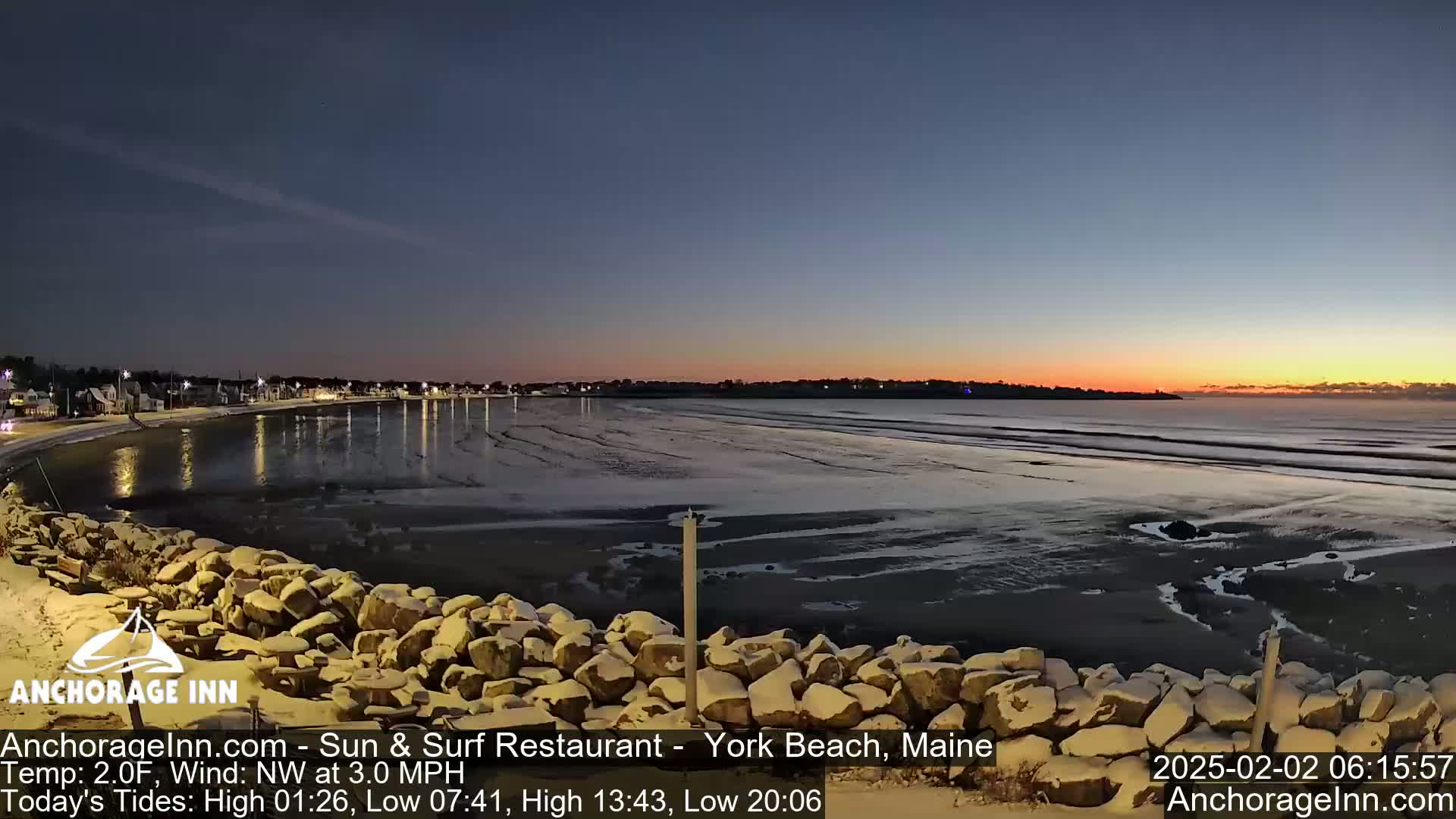 A snow-dusted rocky shoreline overlooks a calm, dark ocean at sunrise, with a town visible in the distance.