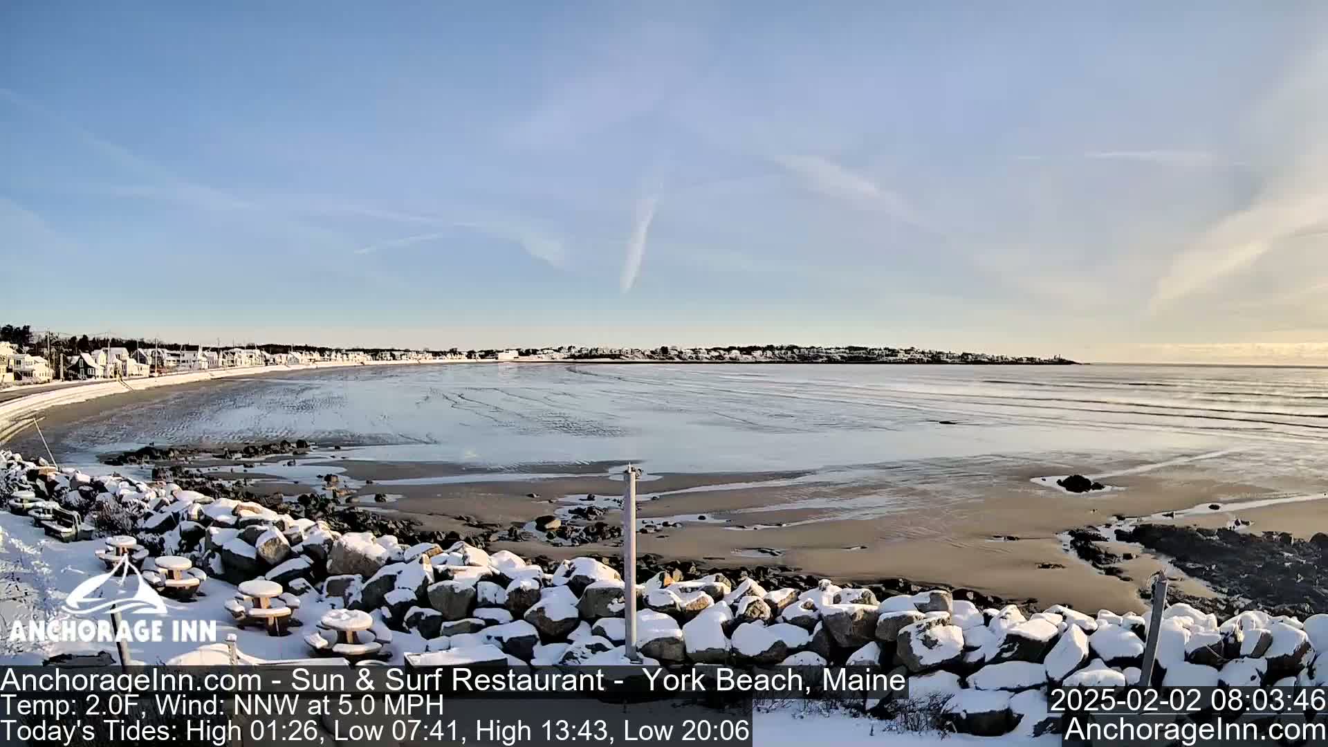 A snow-covered rocky shoreline overlooks a partially frozen beach and calm ocean under a clear, sunny sky.