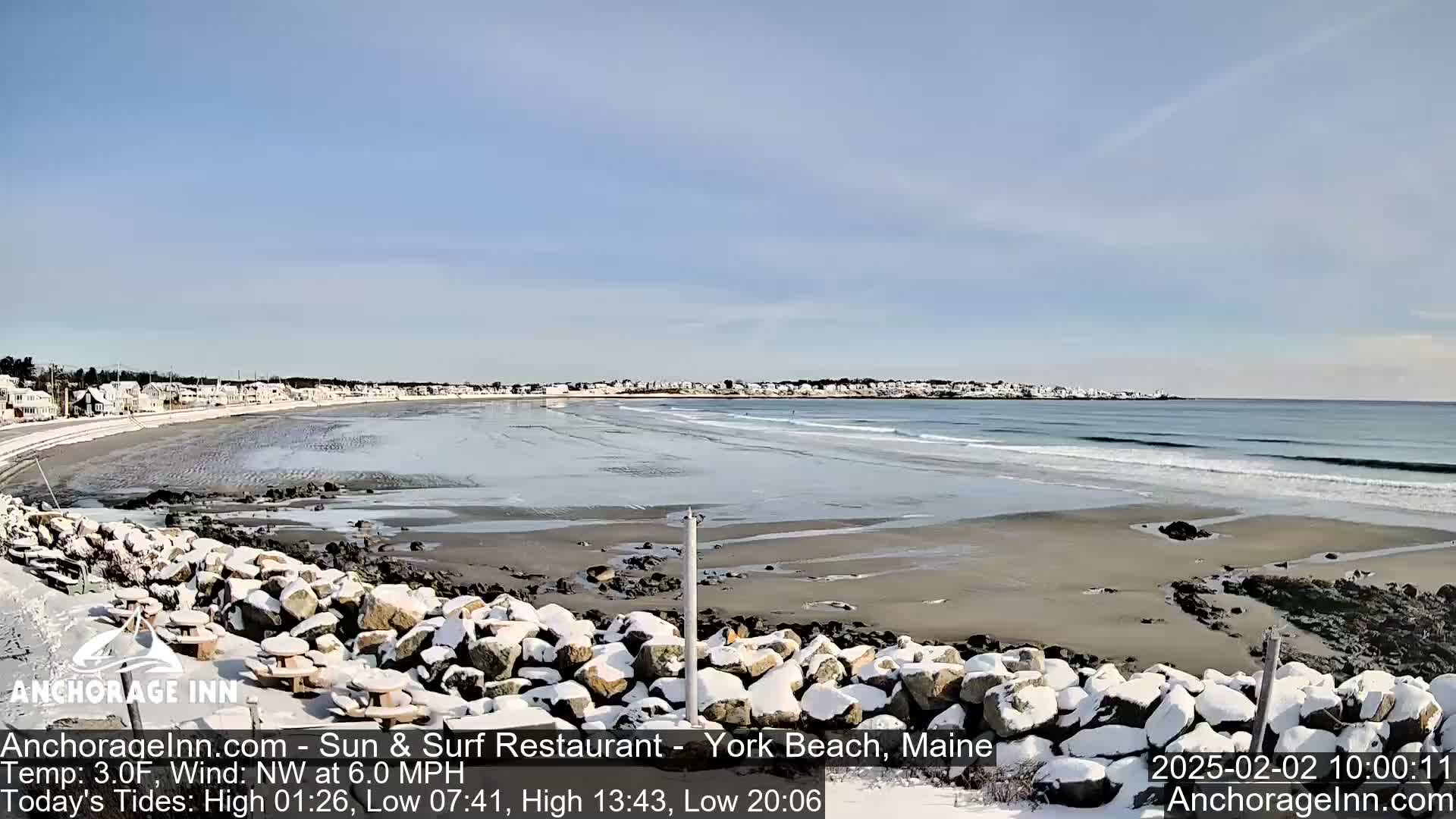 A snow-covered rocky shoreline overlooks a sandy beach and calm ocean waves under a clear blue sky.