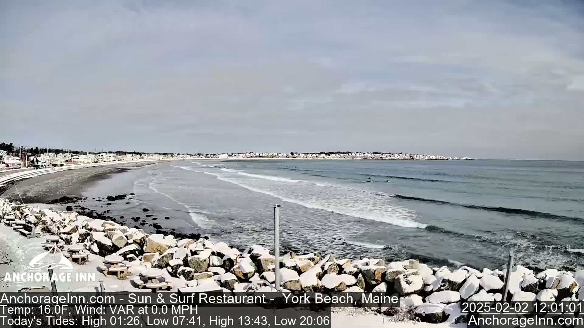 A snow-dusted rocky shoreline overlooks a calm ocean with small waves and a few surfers in the distance, under a mostly cloudy sky.
