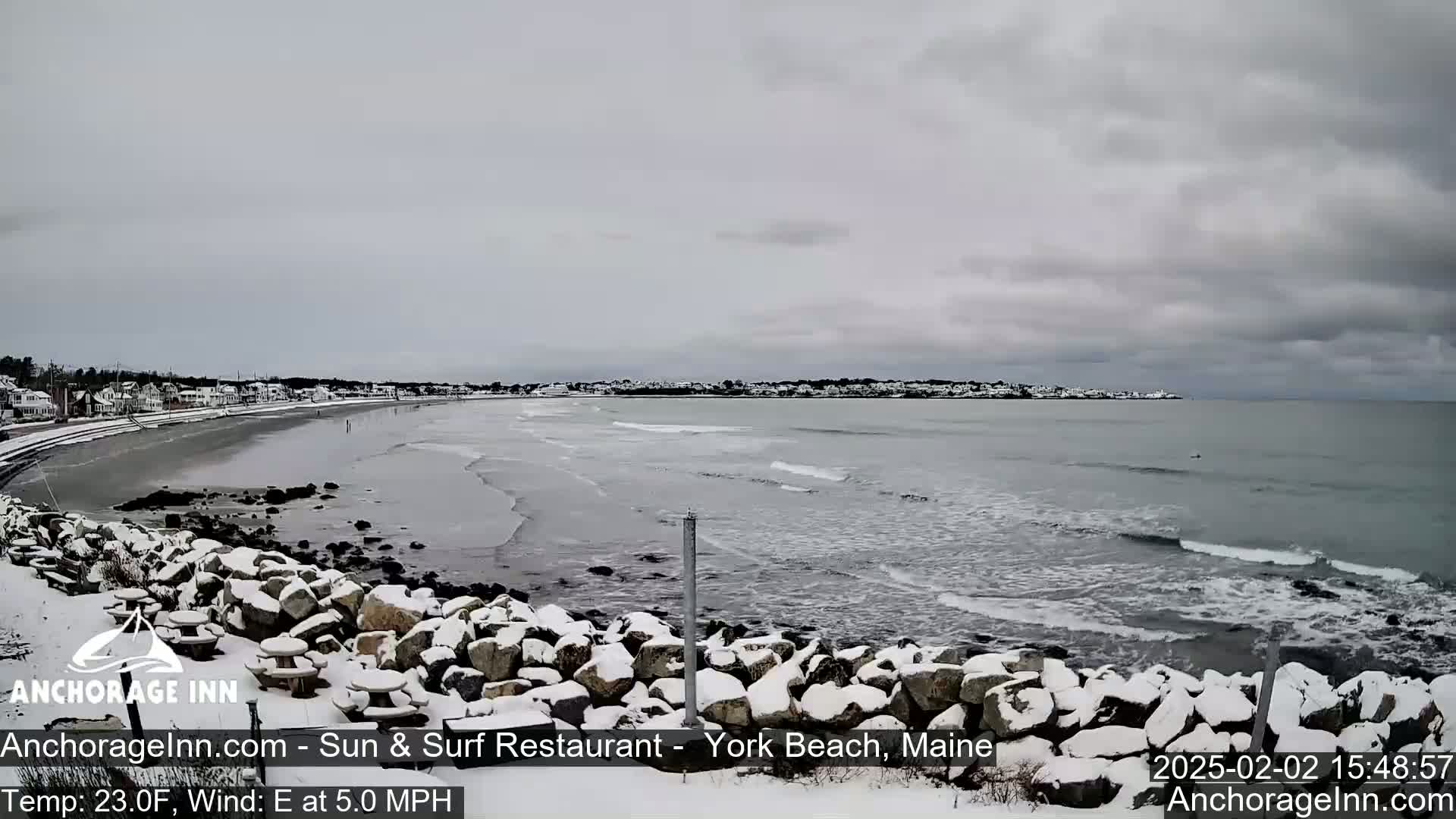 A snow-covered rocky shoreline overlooks a gray, wavy ocean with a town visible in the distance under an overcast sky.
