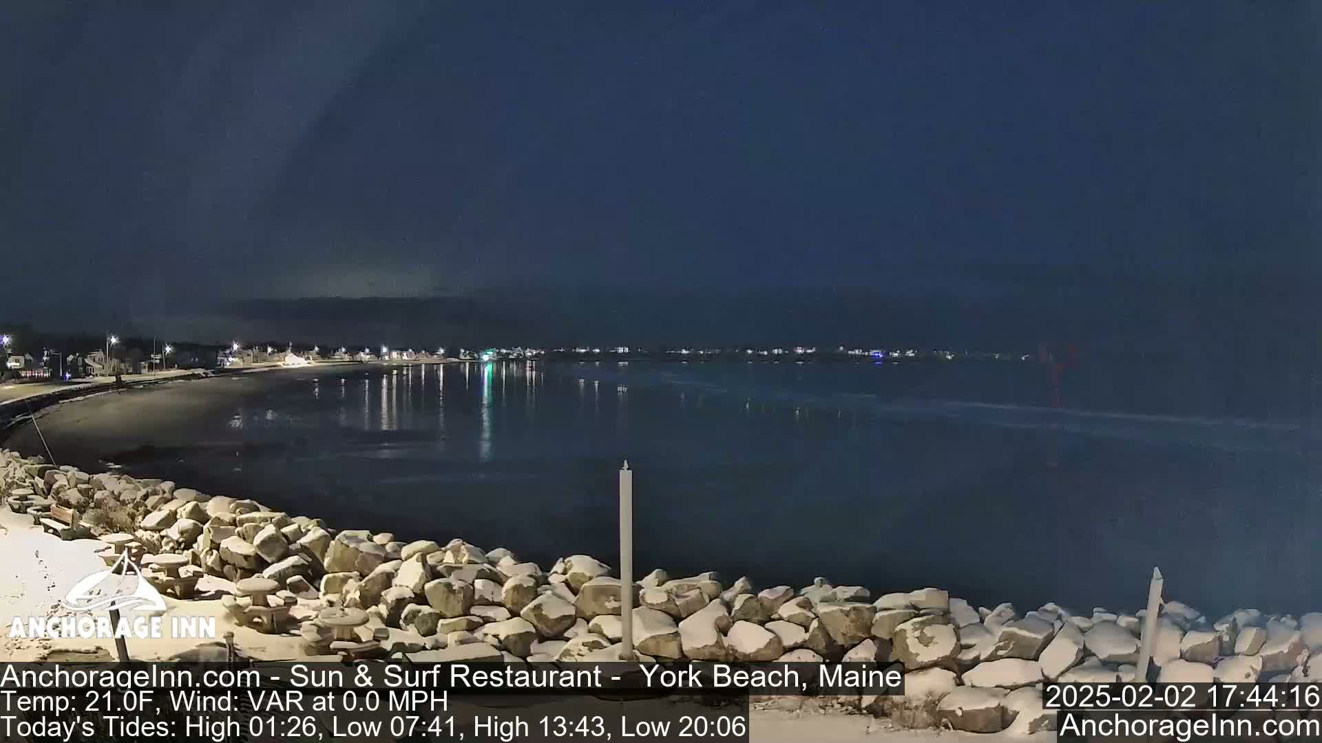 A nighttime view of a snow-dusted rocky shoreline and calm bay, with a town's lights visible in the distance under a dark sky.