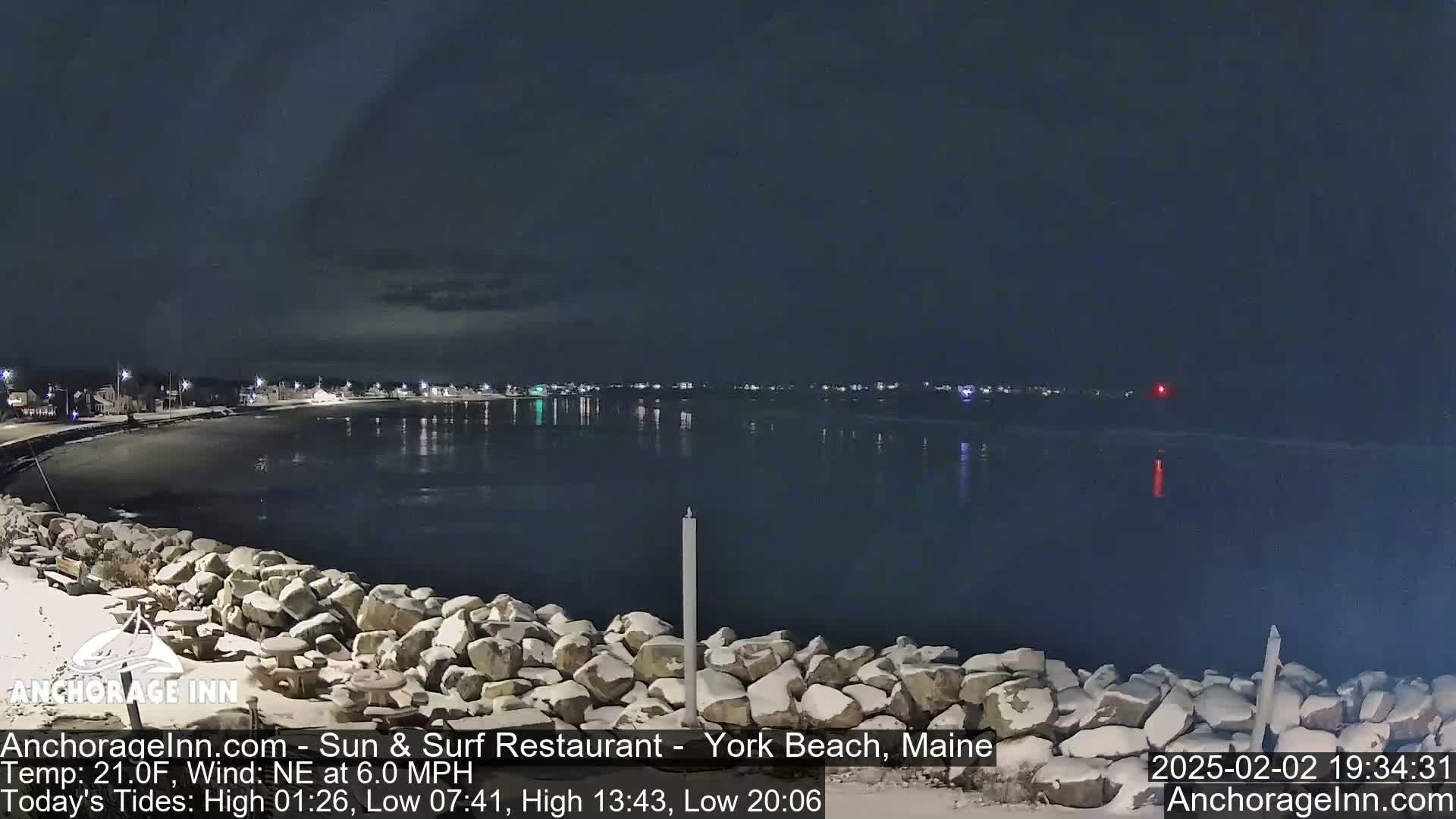 A nighttime view of a snow-dusted rocky shoreline and calm, dark water with a town faintly visible in the distance under a mostly clear sky.