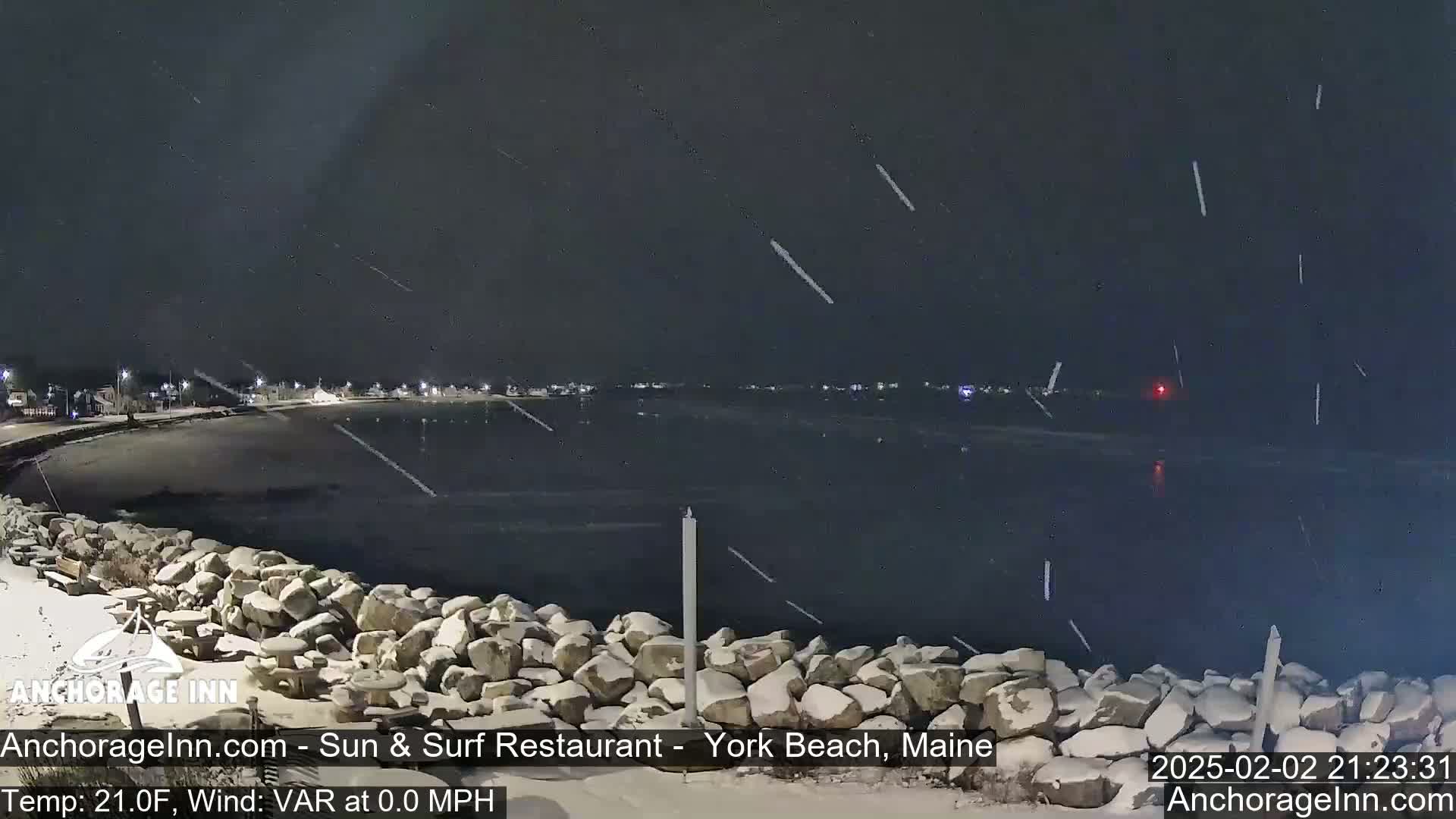 A nighttime view of a snow-covered rocky shoreline and calm, dark water under a lightly snowing sky.
