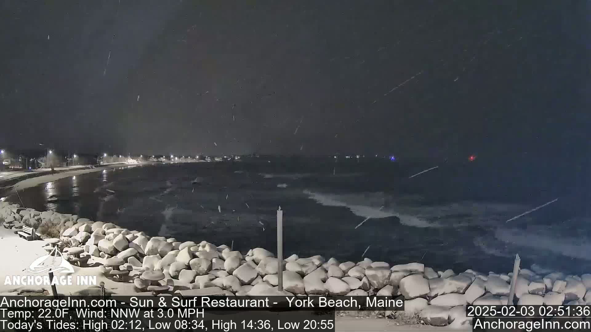 A snow-covered rocky shoreline at night shows a dark, choppy ocean under a lightly falling snow.