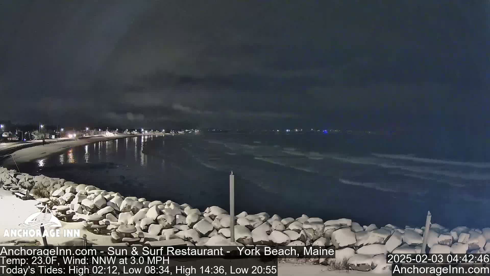 A snow-covered rocky shoreline at night overlooks a dark, gently waving ocean under a cloudy sky.