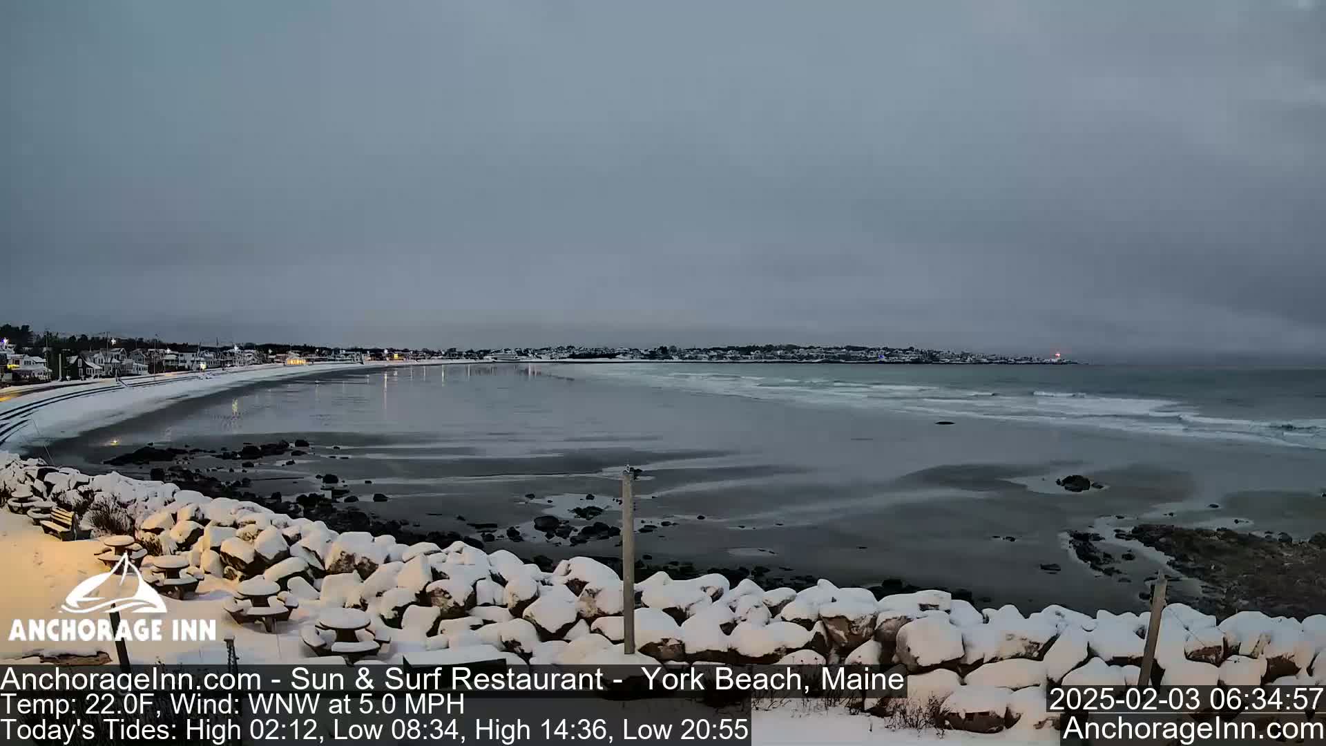 A snow-covered rocky shoreline overlooks a calm, gray ocean with gentle waves under an overcast sky.