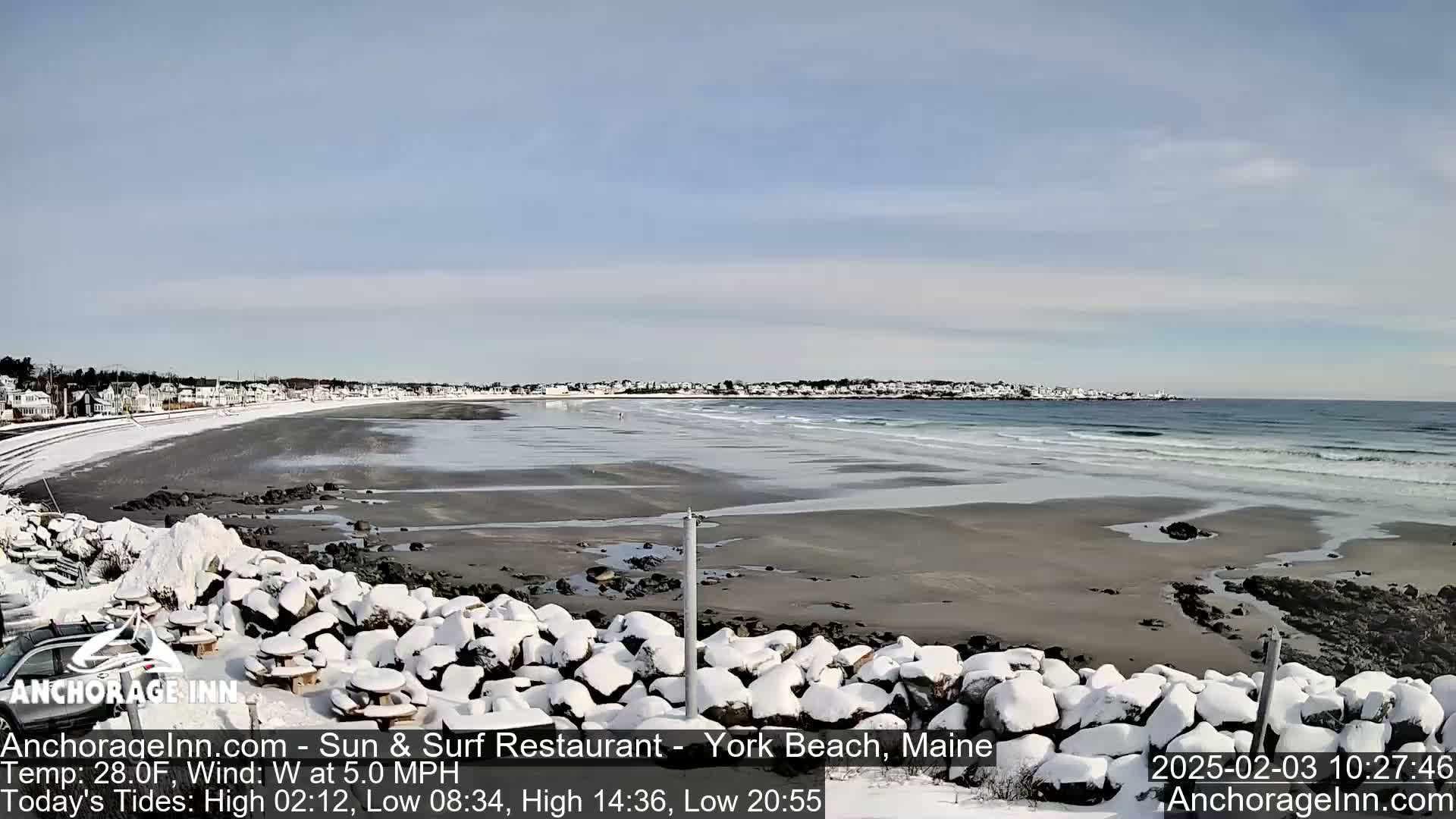 A snow-covered rocky shoreline overlooks a beach with gentle waves and a town in the distance under a partly cloudy sky.