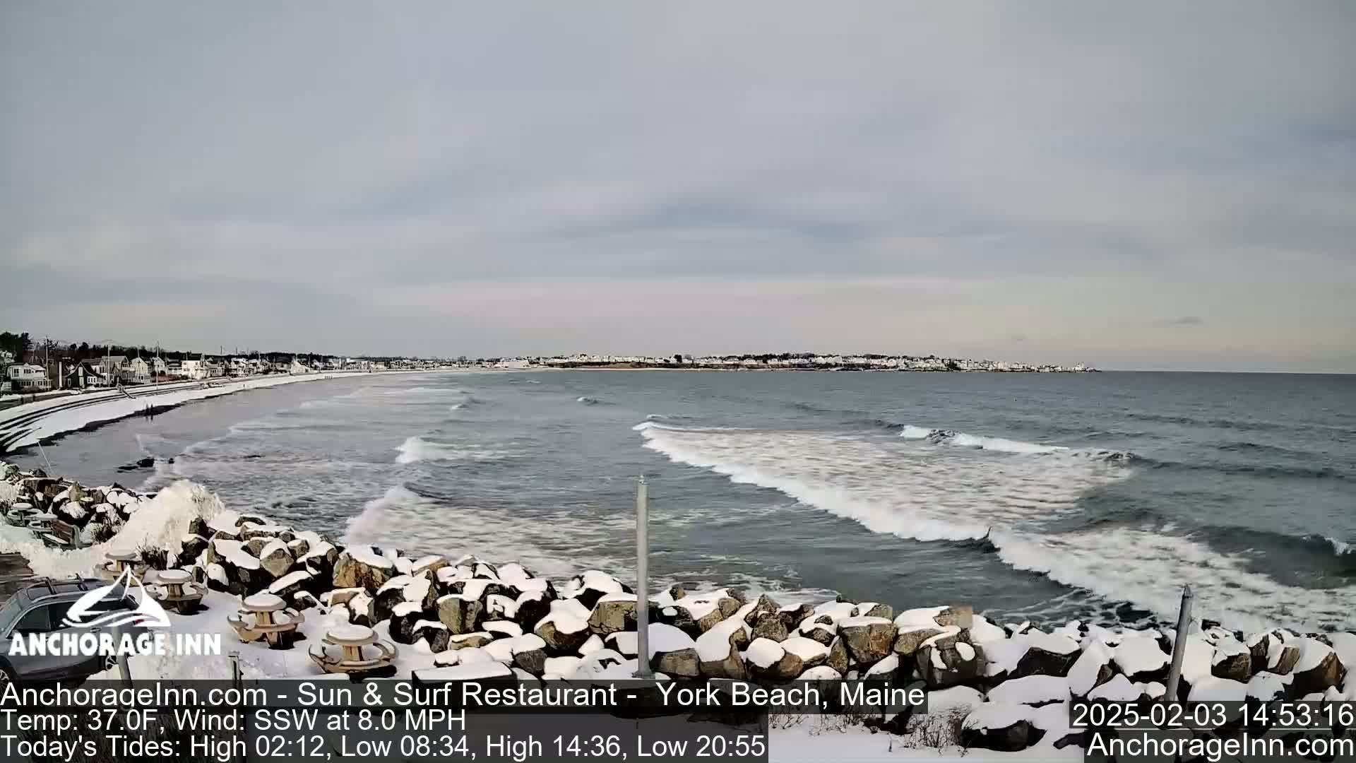 A snow-covered rocky shoreline with small waves rolling onto a beach in front of a town under an overcast sky.