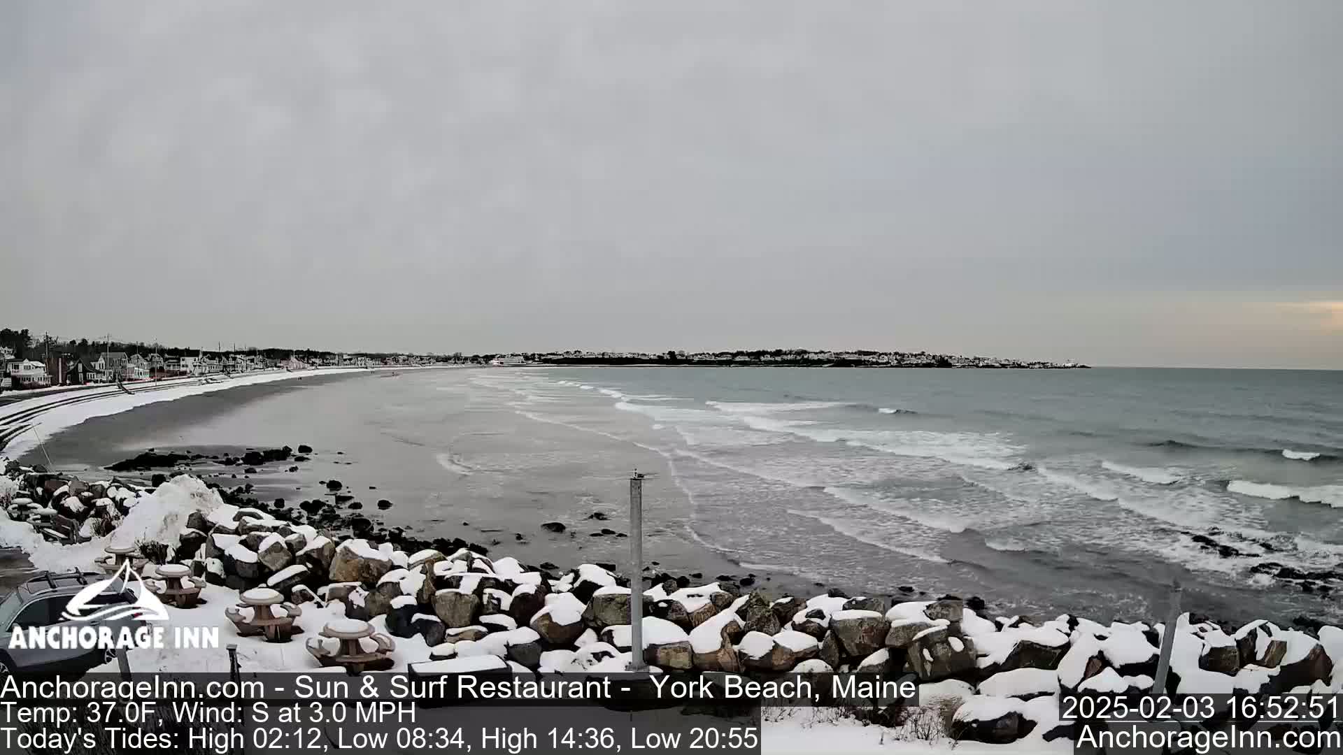 A snow-covered rocky shoreline meets a gently wavy ocean under an overcast sky.