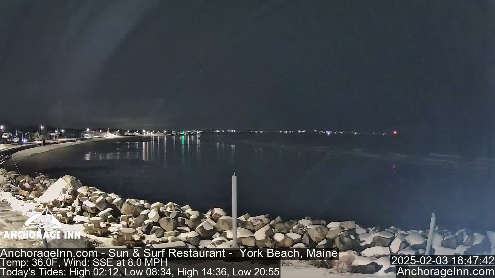 A snow-dusted rocky shoreline at night overlooks a calm, dark body of water reflecting distant lights under a clear, dark sky.