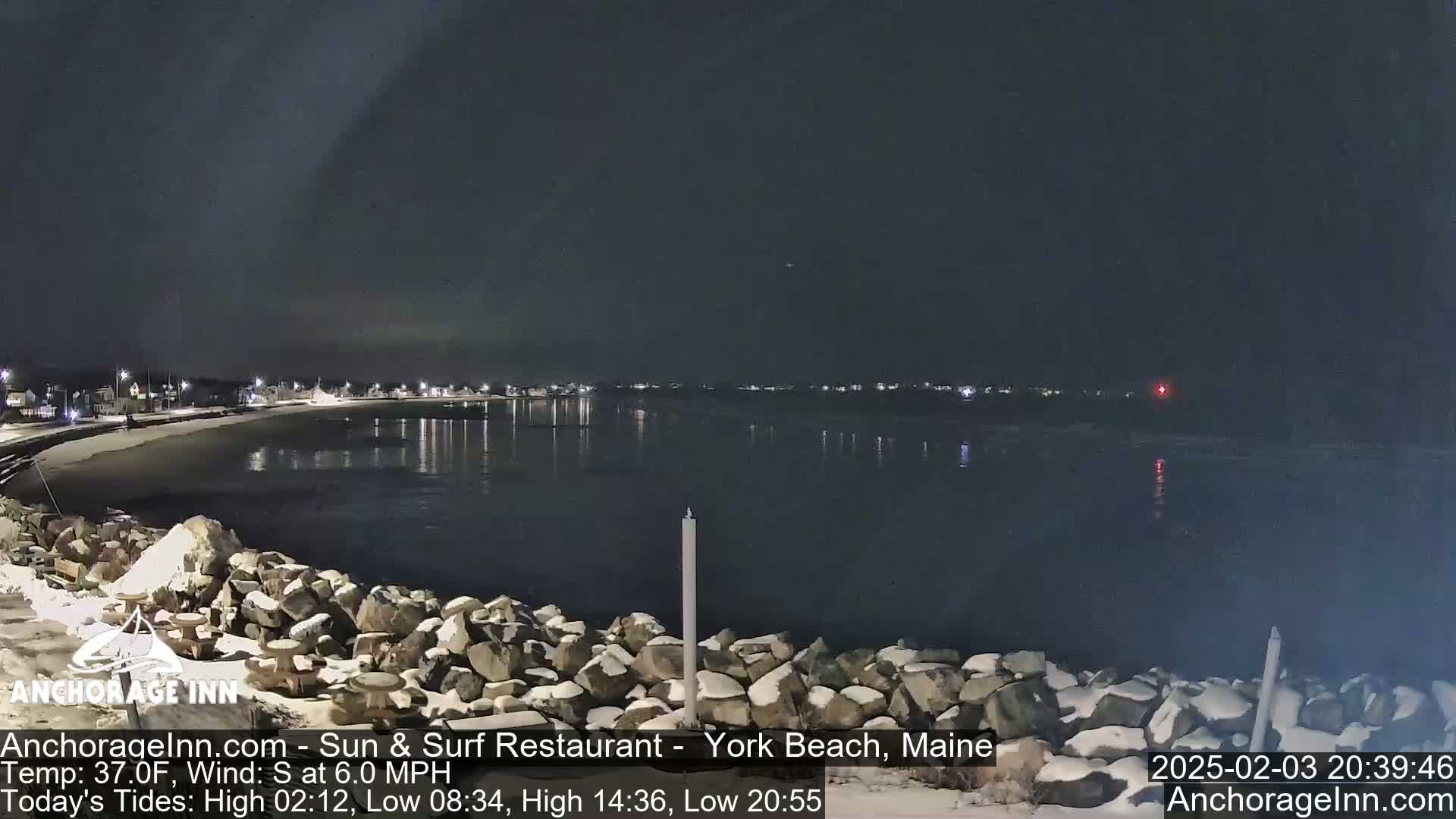 A nighttime view of a snow-dusted rocky shoreline and calm water, with a town faintly lit in the distance under a clear, dark sky.