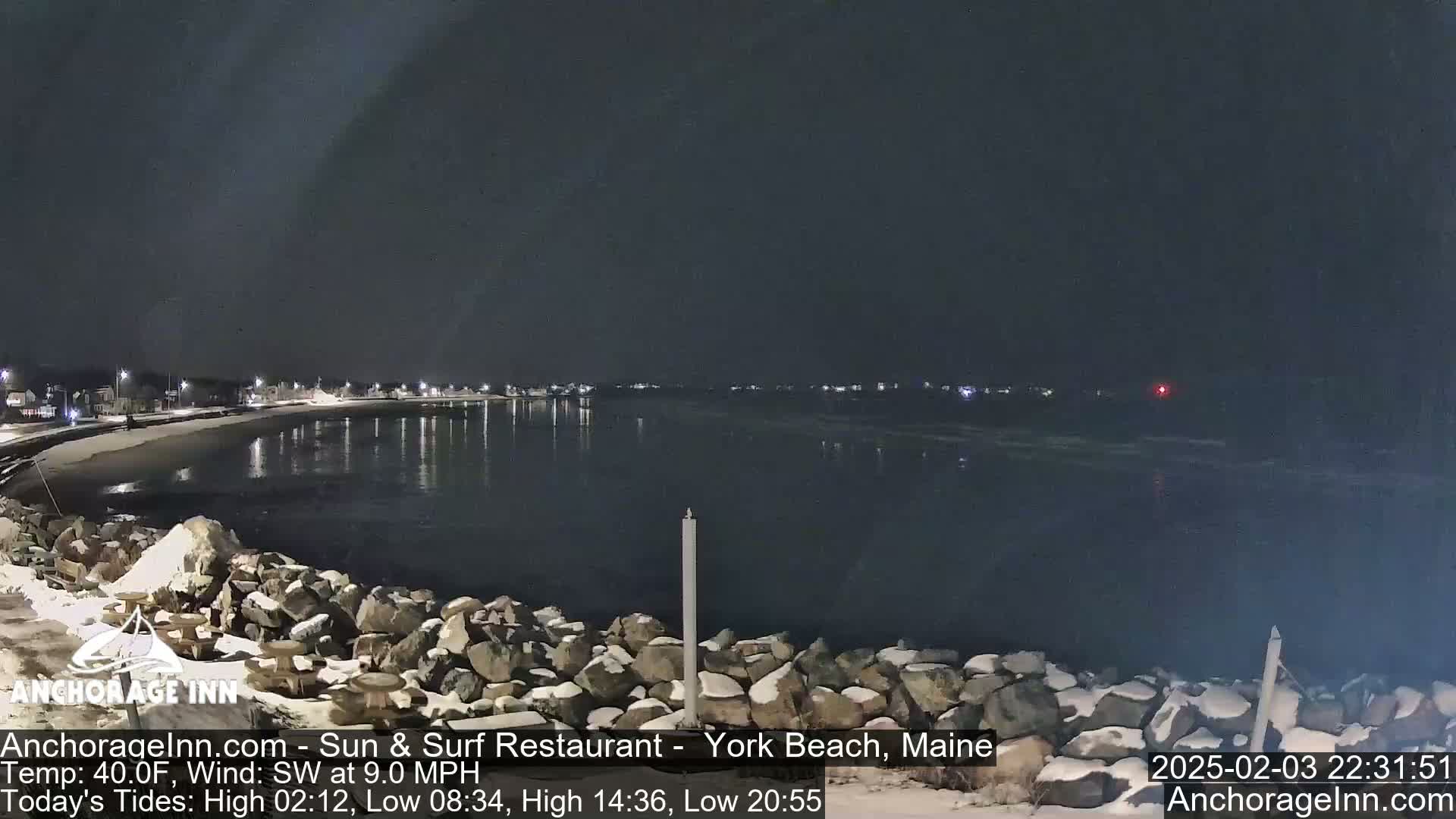 A nighttime view of a snow-dusted rocky shoreline and calm water, with lights visible across the water and along the coast under a dark sky.