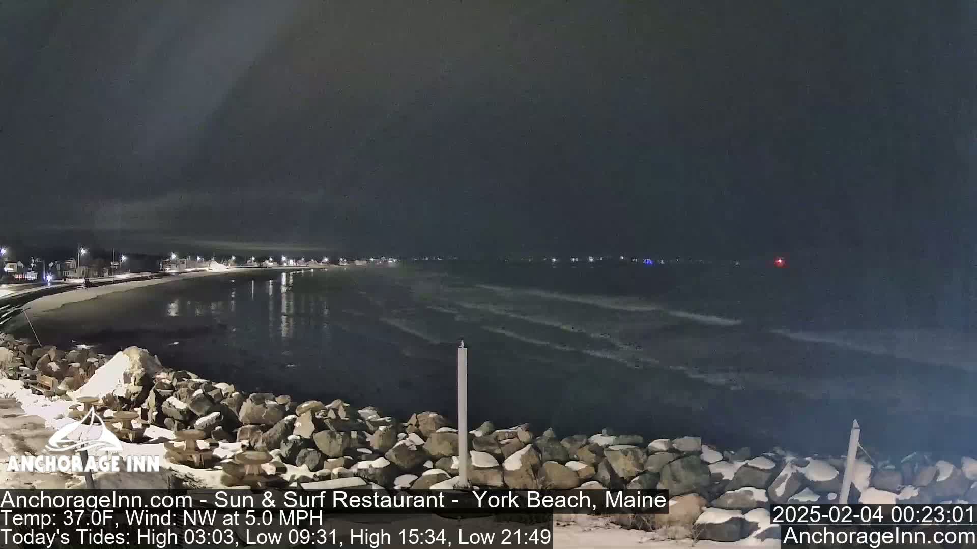 A nighttime view of a dark, wavy ocean next to a snow-covered rocky shoreline with faintly lit buildings in the distance.