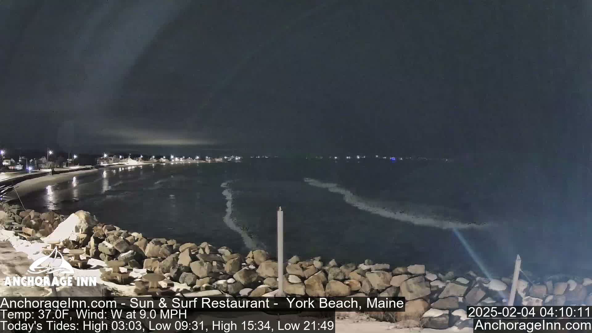 A nighttime view of a dark, wavy ocean meeting a rocky shoreline under a mostly clear sky.