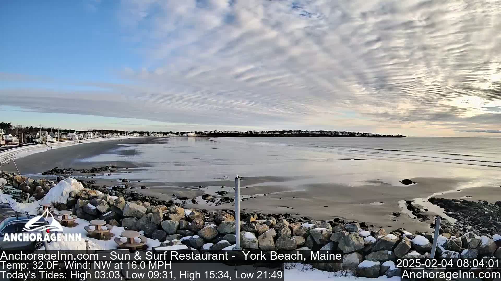 A partially snowy beach with a calm ocean and a town visible in the distance under a mostly cloudy sky.