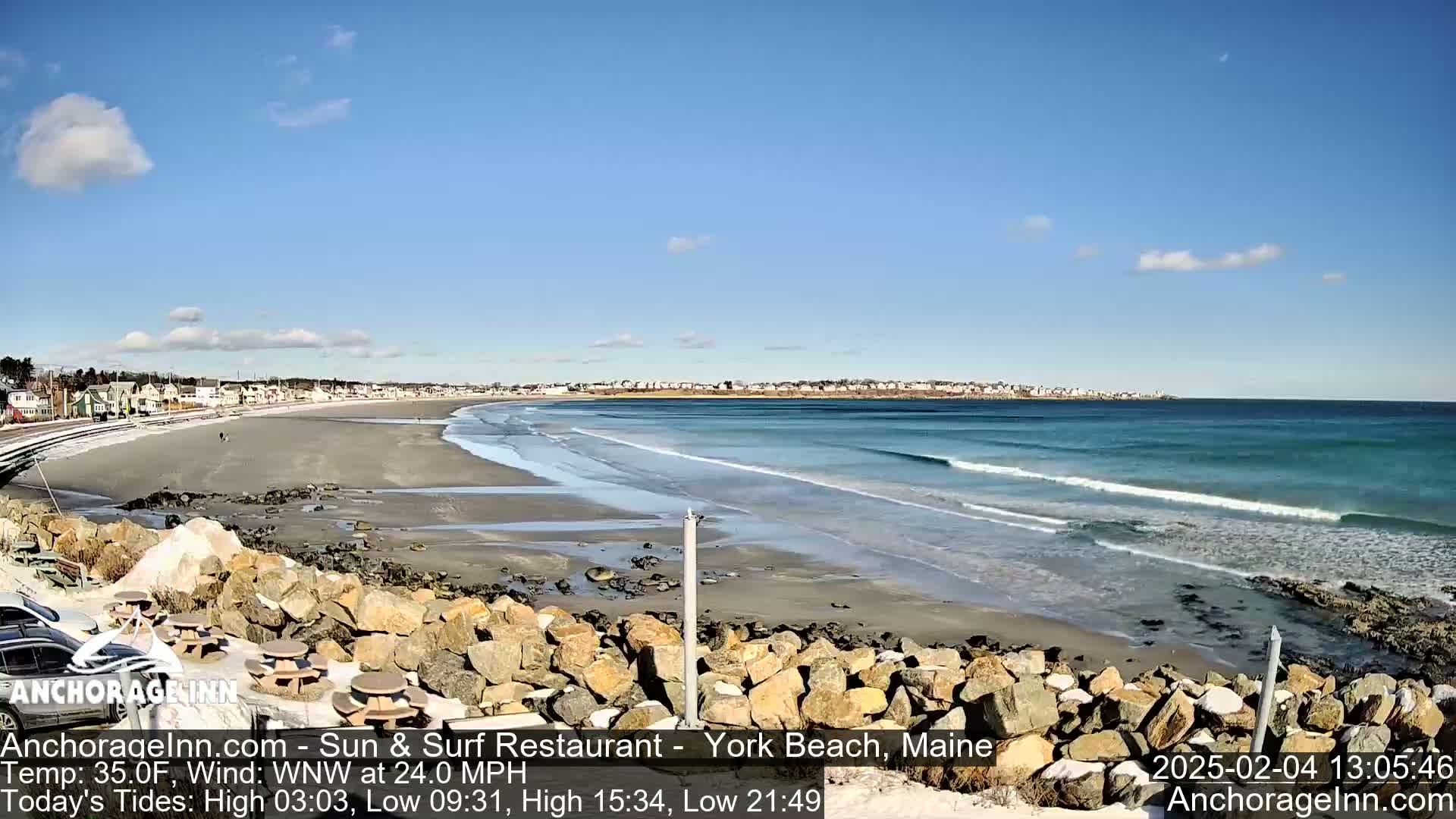 A sandy beach with a rocky shoreline, gentle waves, and a town in the distance under a mostly clear, sunny sky.