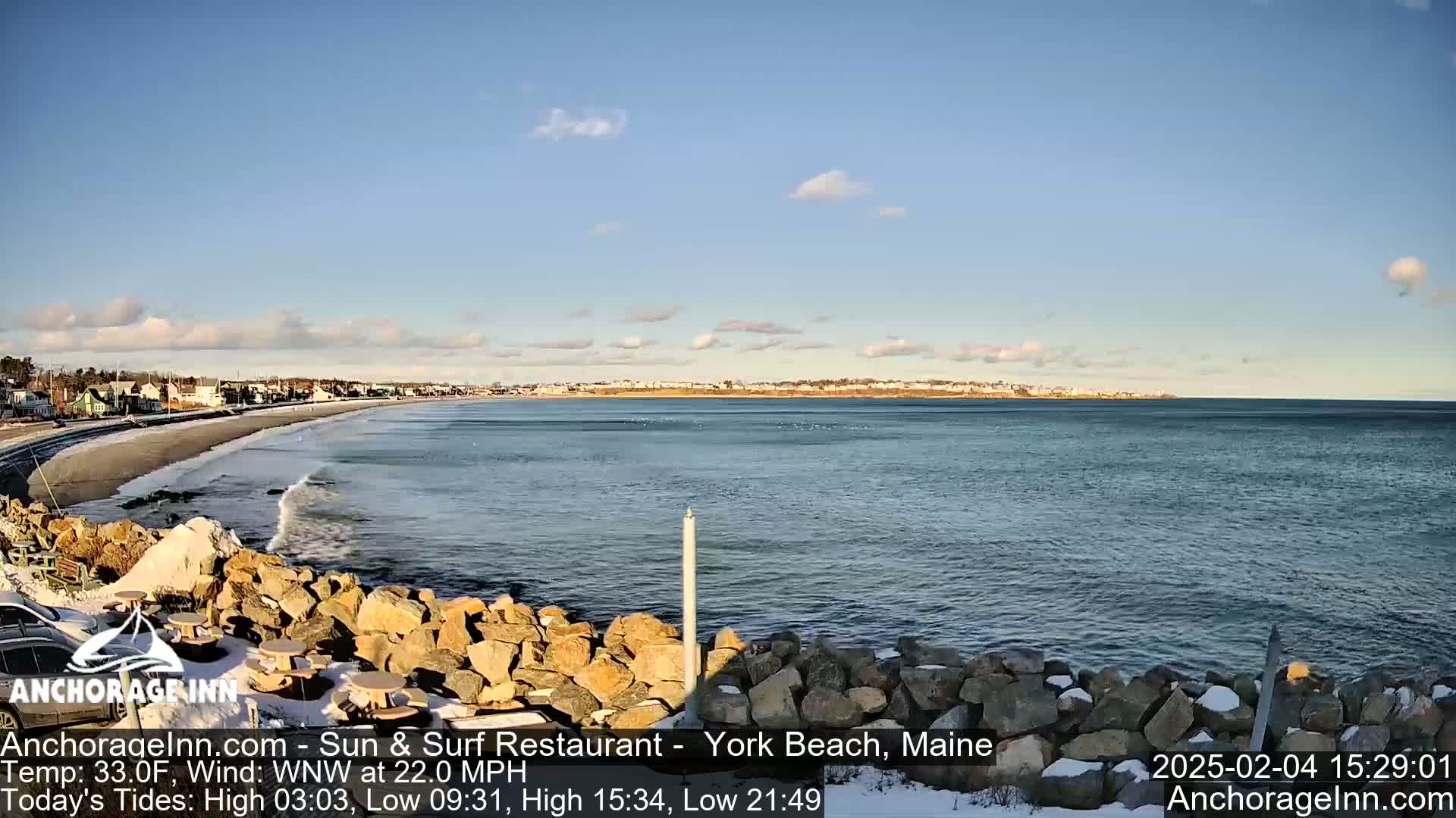A rocky shoreline with a partially snow-covered beach and calm ocean is visible under a partly cloudy sky.