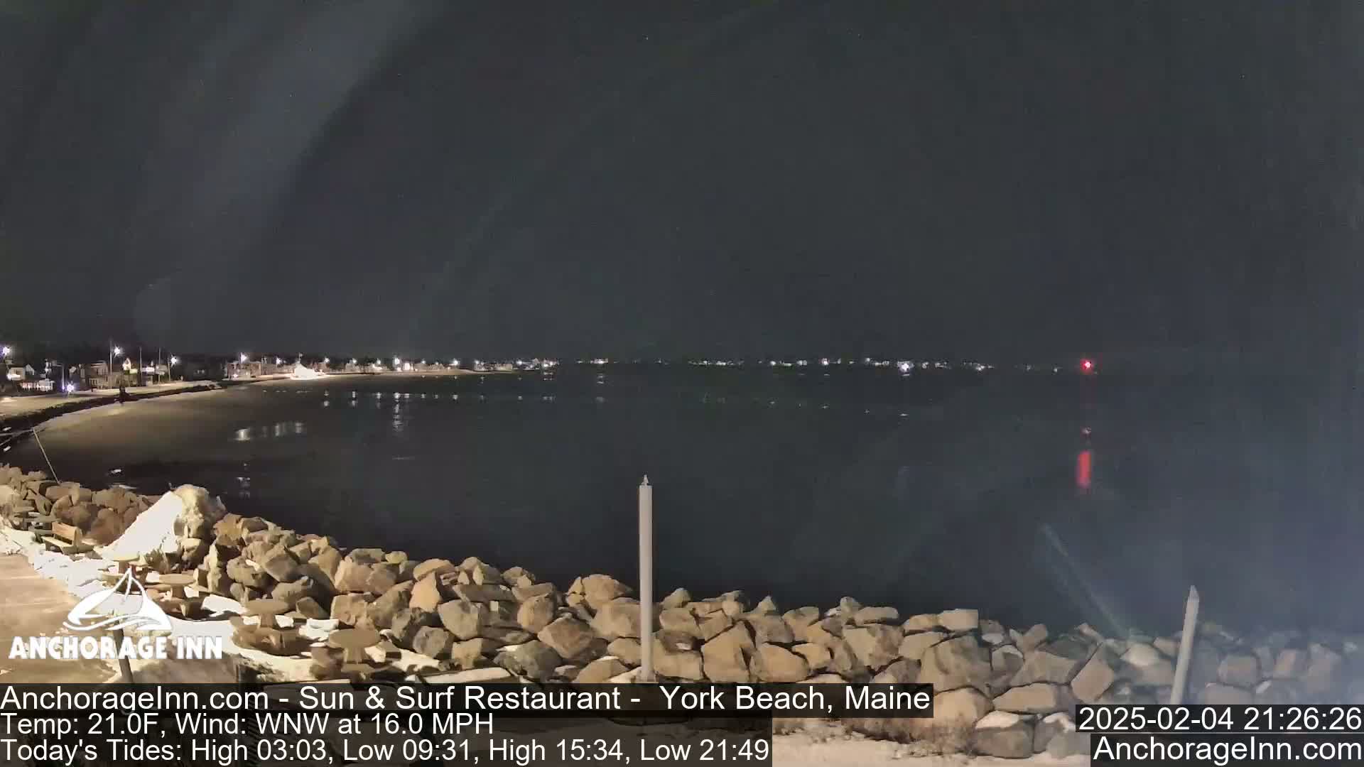 A nighttime coastal scene shows a dark, calm body of water reflecting the lights of a distant town under a clear, dark sky.