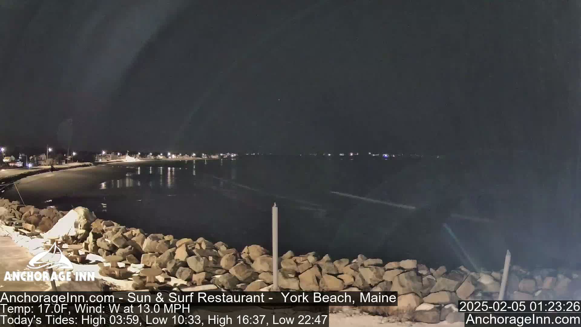 A nighttime view of a rocky shoreline and calm dark water, with a town faintly visible in the distance under a clear sky.