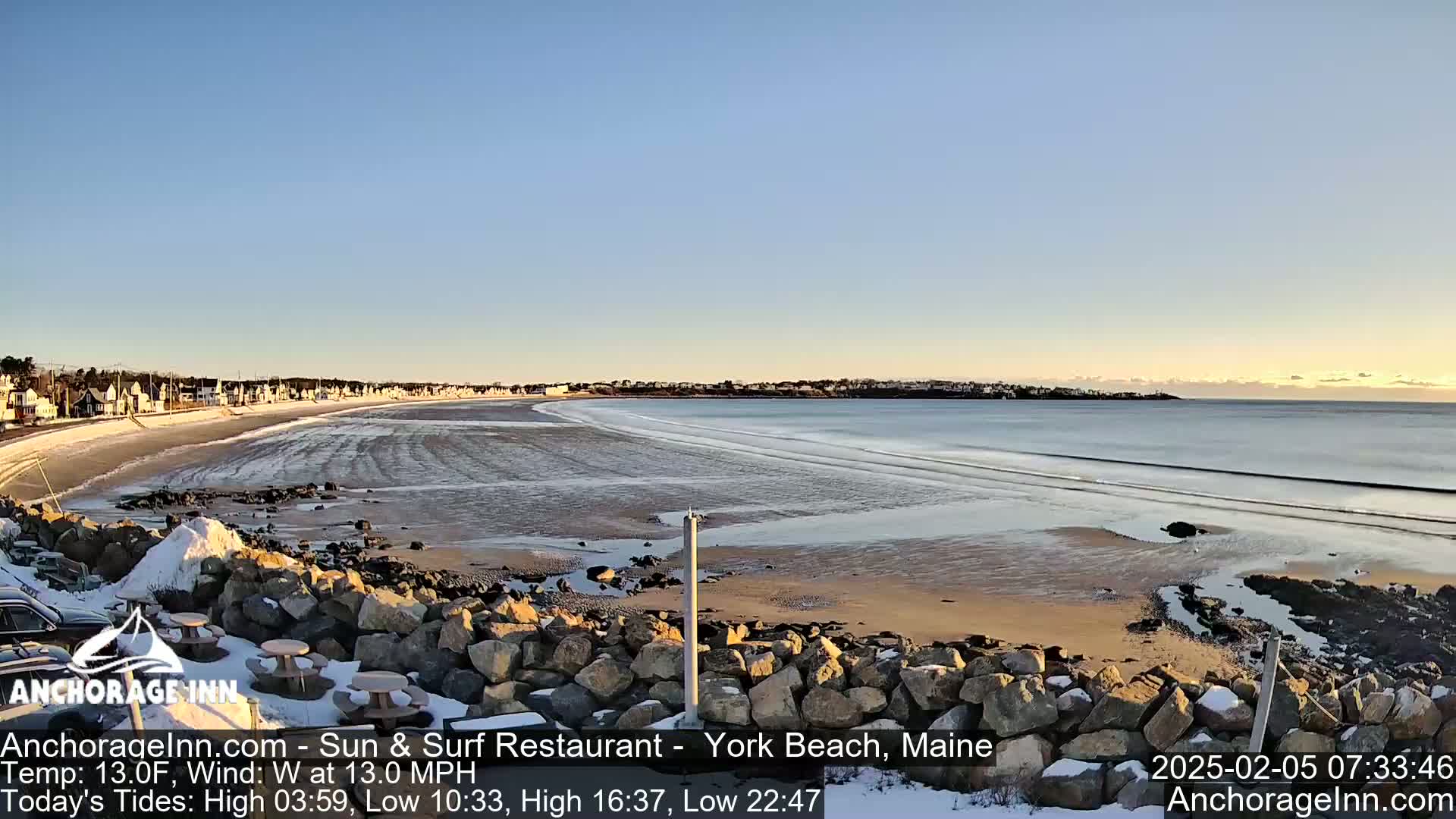 A partly frozen beach with a row of houses in the distance under a clear blue sky.