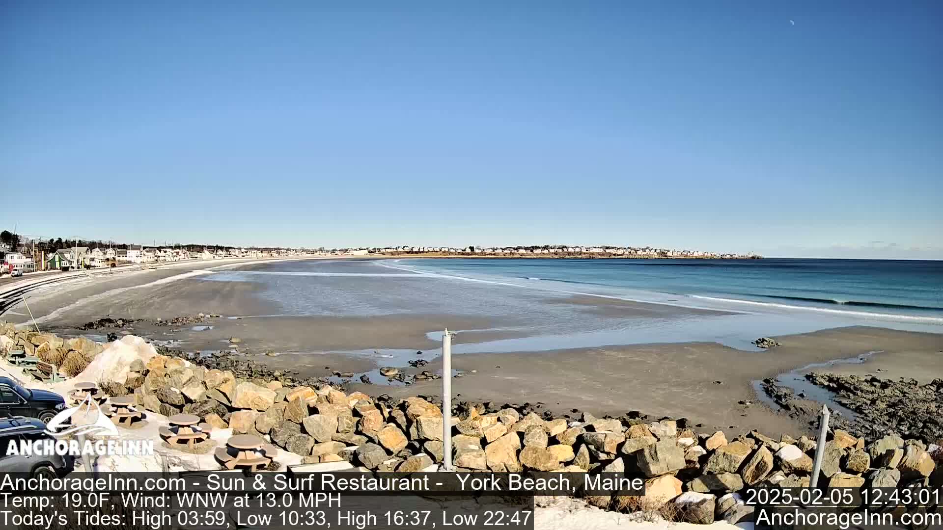 A sandy beach with a calm ocean stretches out under a clear blue sky, with a rocky outcrop in the foreground and buildings visible in the distance.