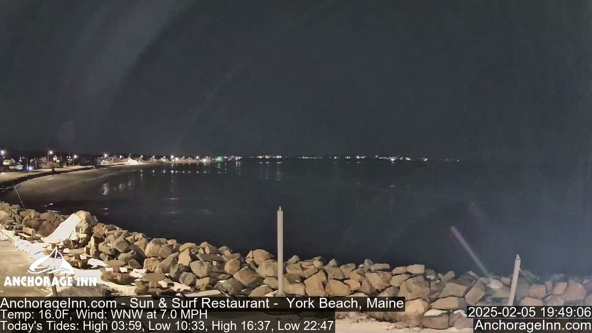 A nighttime coastal scene shows a dark, calm body of water next to a rocky shoreline with distant lights on the opposite shore under a clear, dark sky.