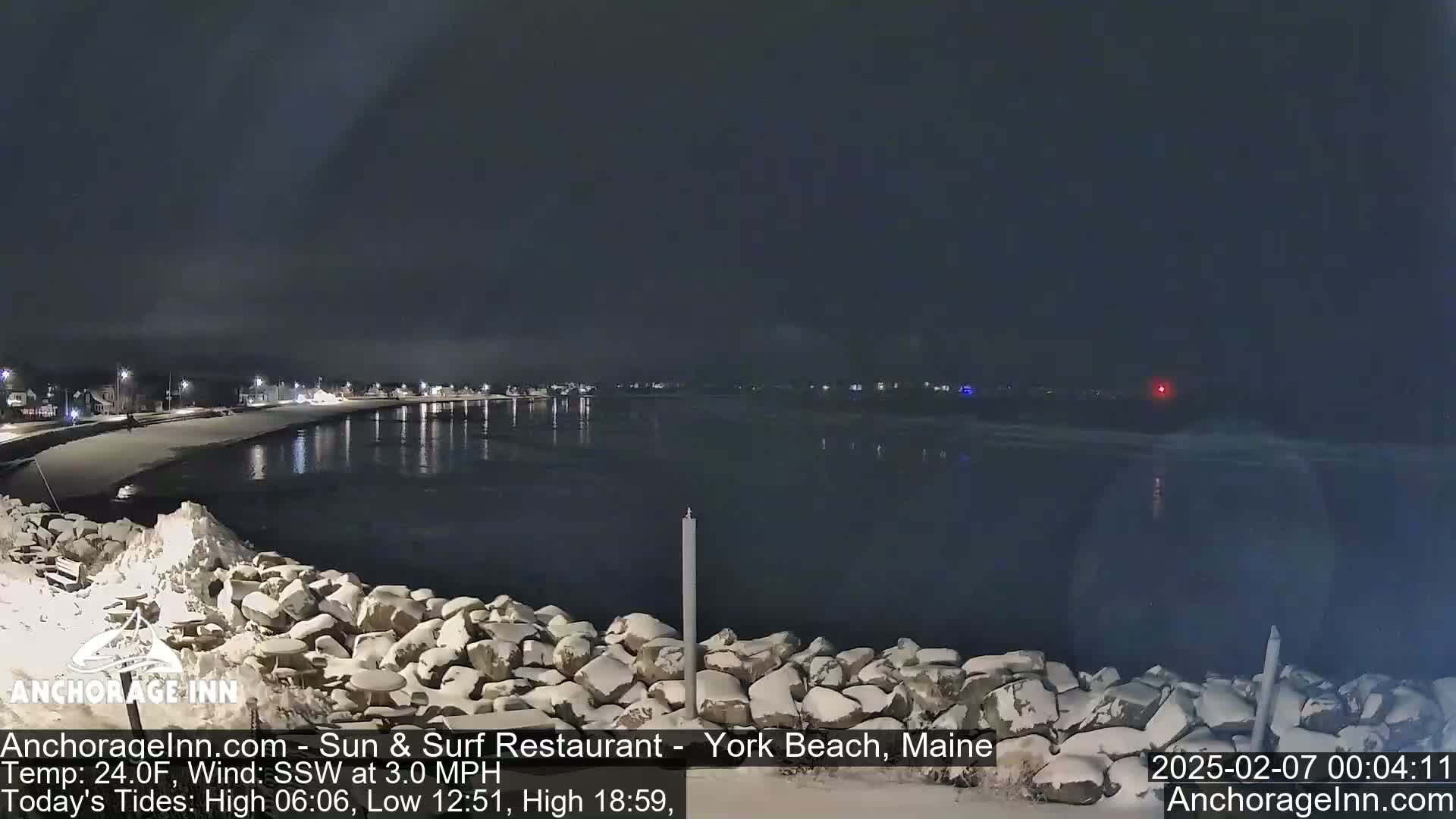 A snow-covered rocky shoreline at night overlooks a calm, dark body of water under a clear, dark sky.